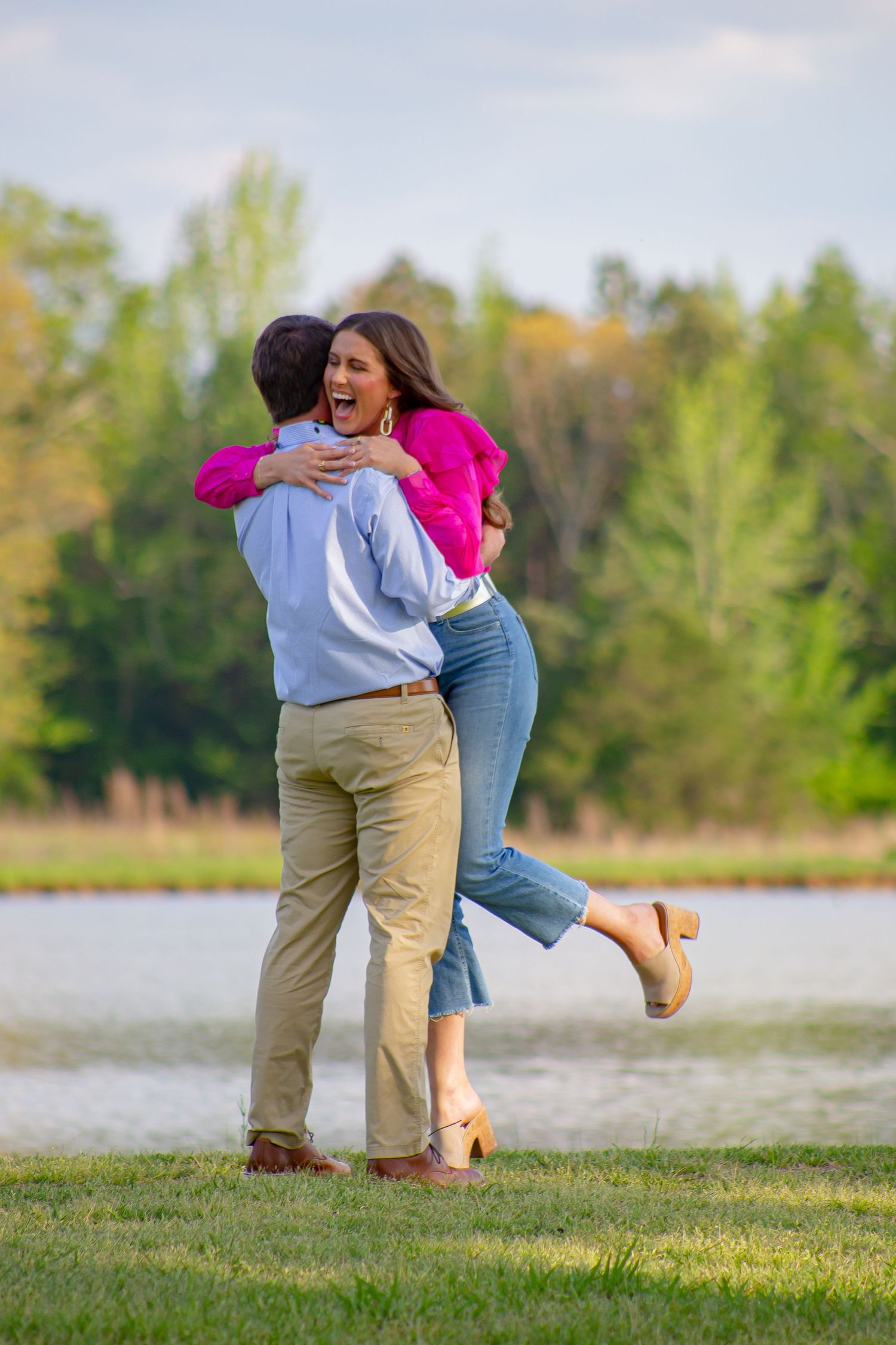 A couple embracing after a proposal on a grassy field with trees in the background.