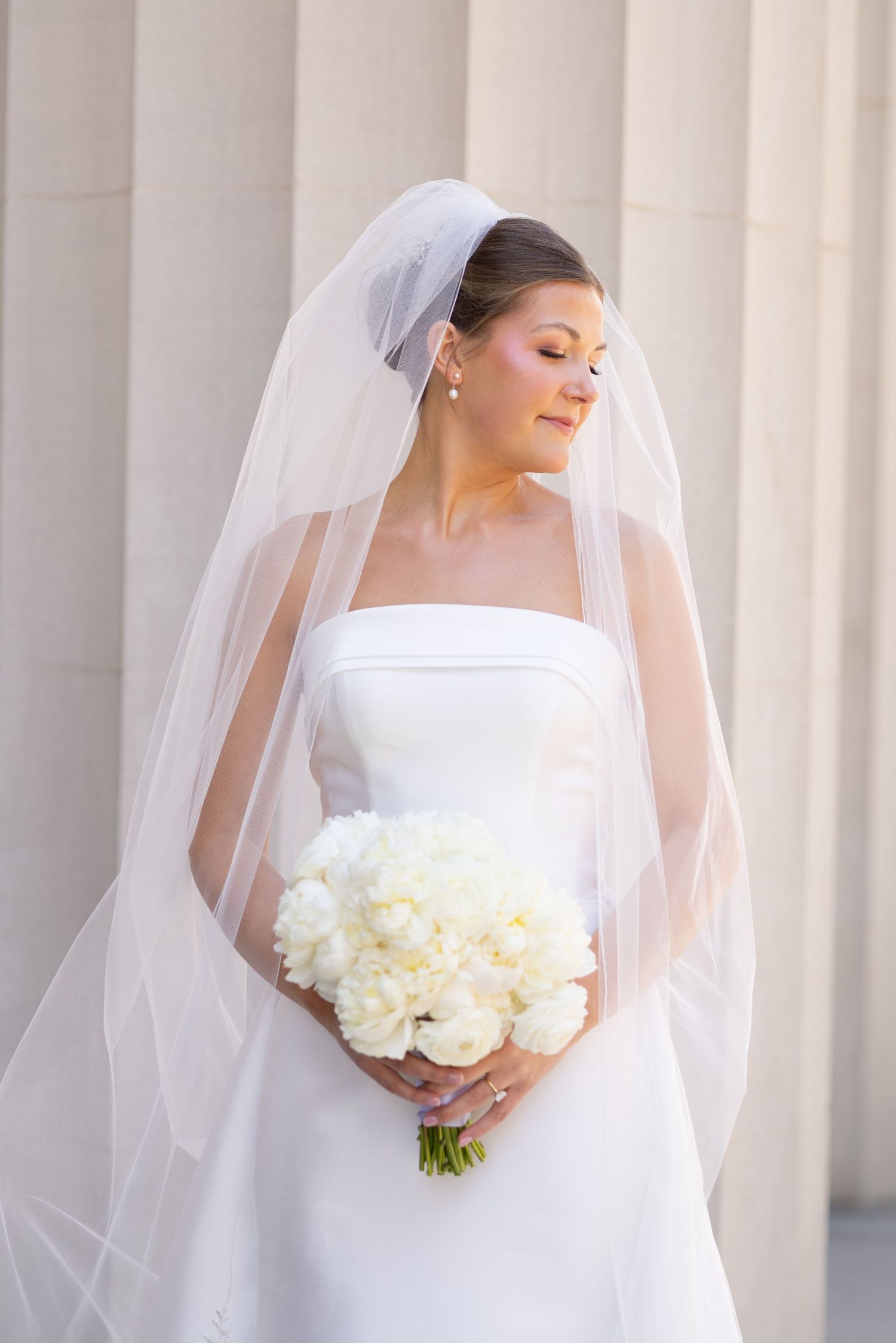 Elegant bride in strapless white gown and veil holds white peony bouquet, posing gracefully beside classic stone columns.