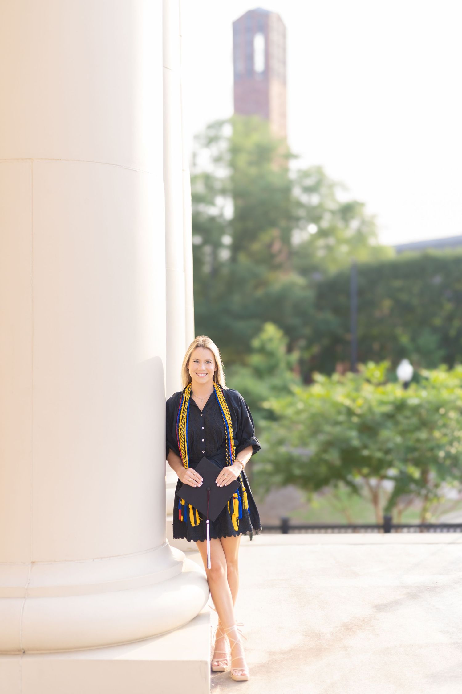 Graduate in black dress and graduation stole stands between white columns on campus.