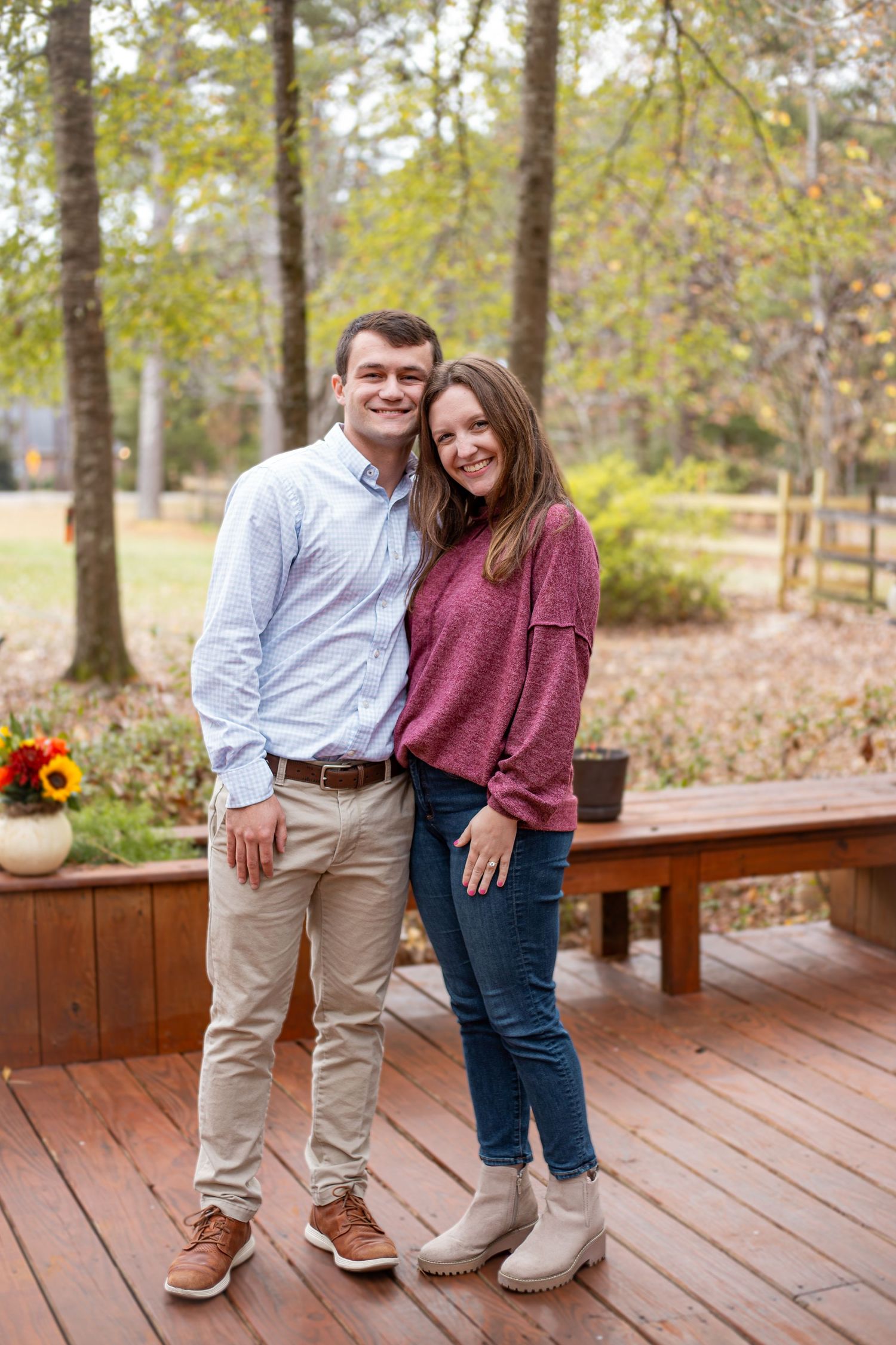 A couple poses together on a wooden deck with autumn trees and sunflowers in the background.