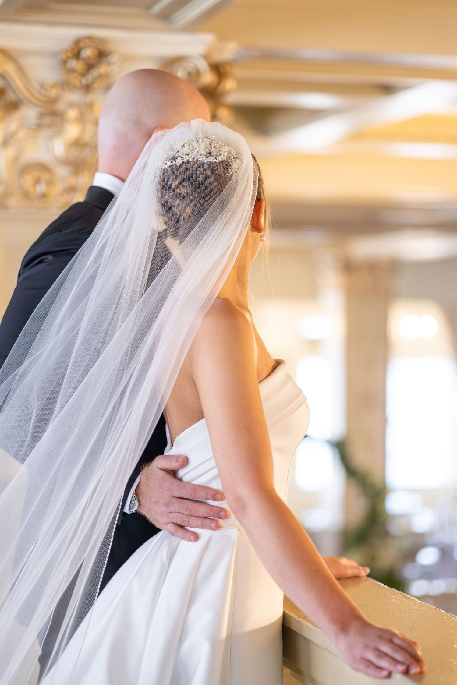 Bride and groom embracing on a balcony, bride wearing a white gown and veil with floral hairpiece in elegant venue.