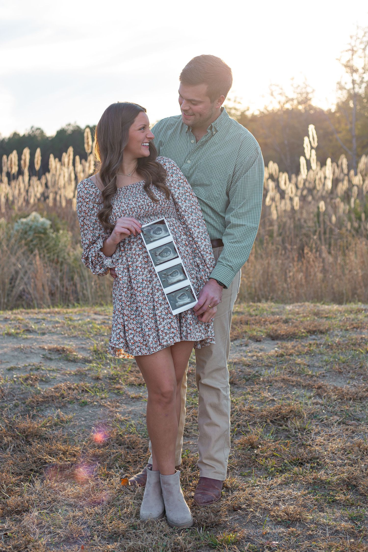 Couple shares intimate moment while holding ultrasound photos in sunlit field.