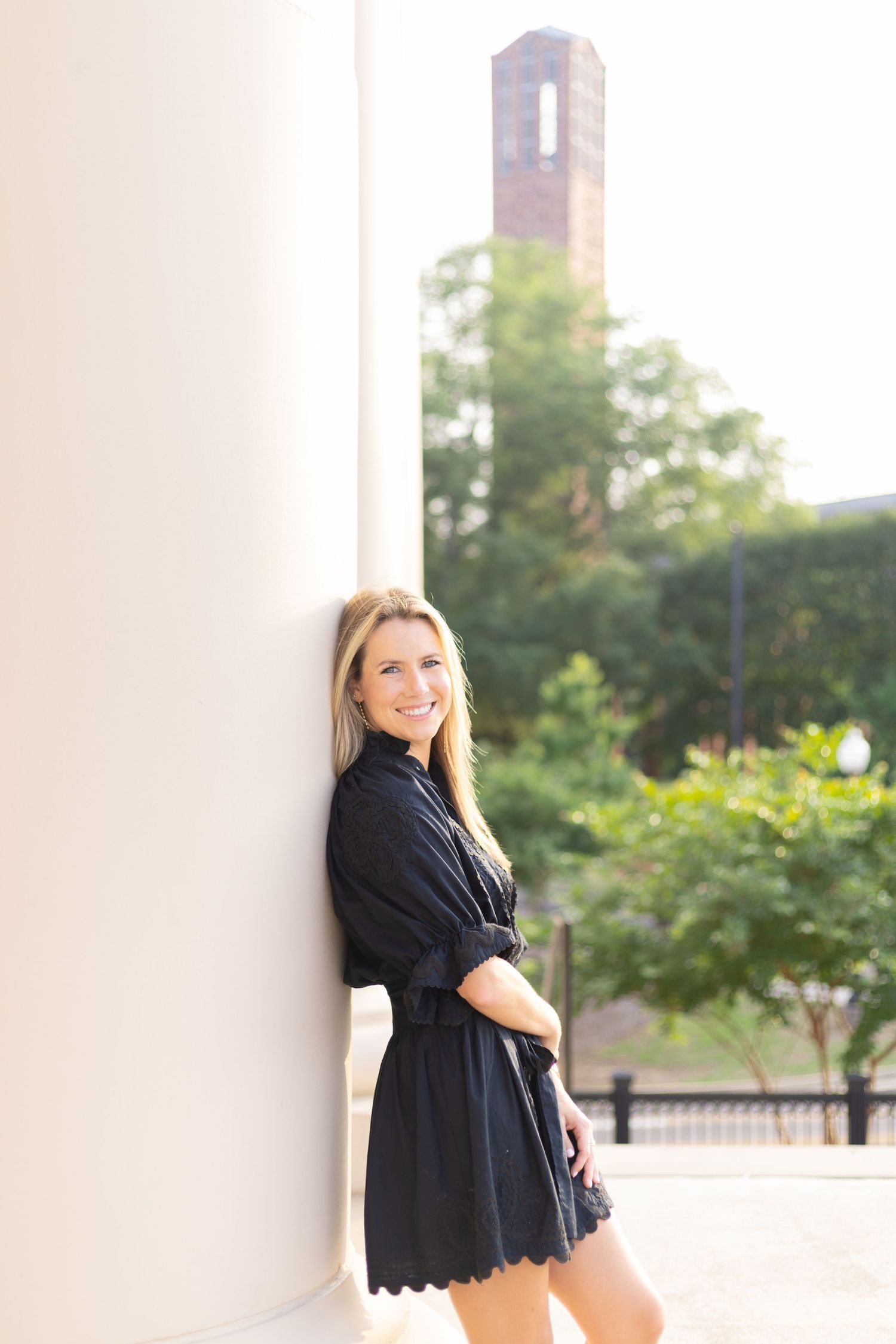 Graduate in black dress leans against white column during outdoor photoshoot.