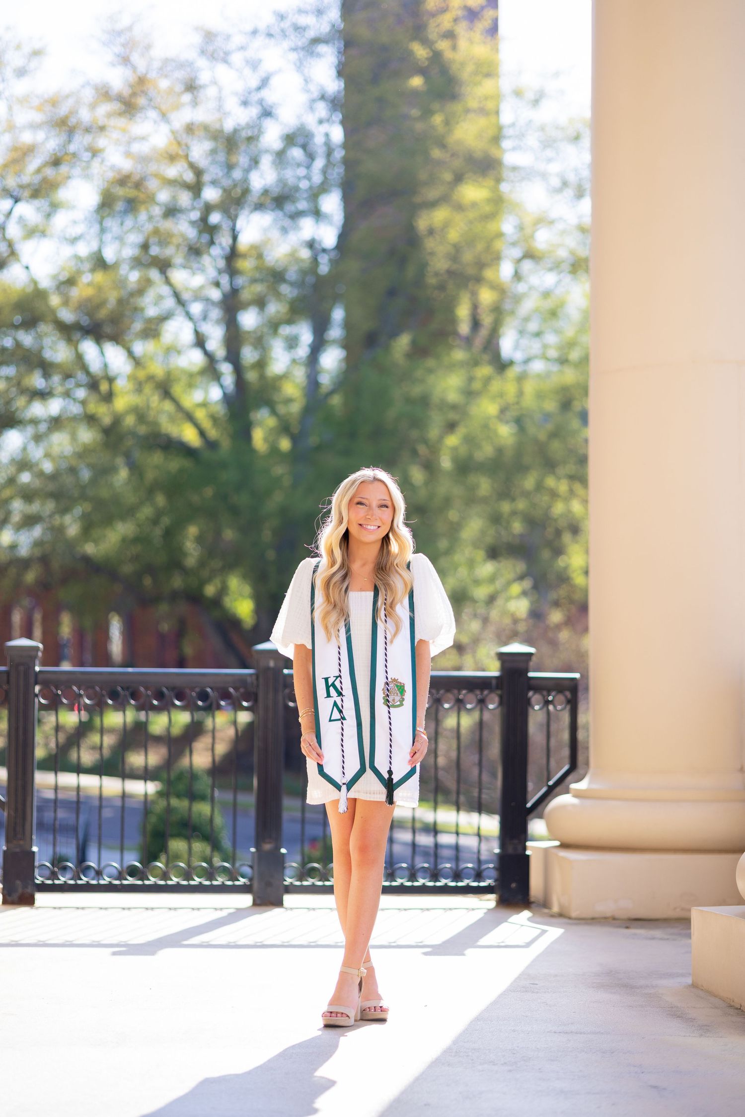 Smiling blonde woman wearing Kappa Delta graduation stole in white dress, posing outdoors near columns and trees.