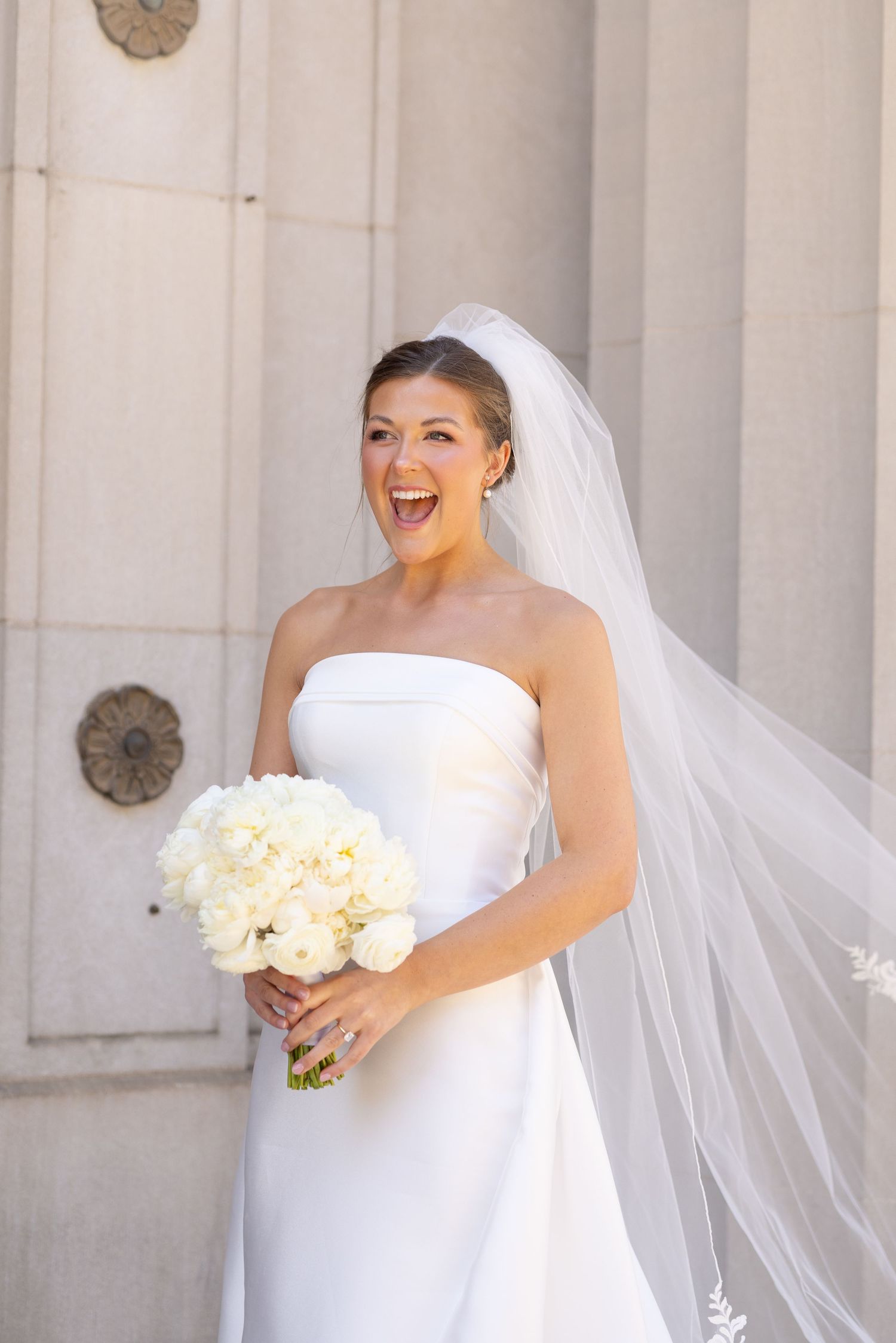 Joyful bride in strapless white gown and flowing veil holds white bouquet, laughing outdoors near stone columns.