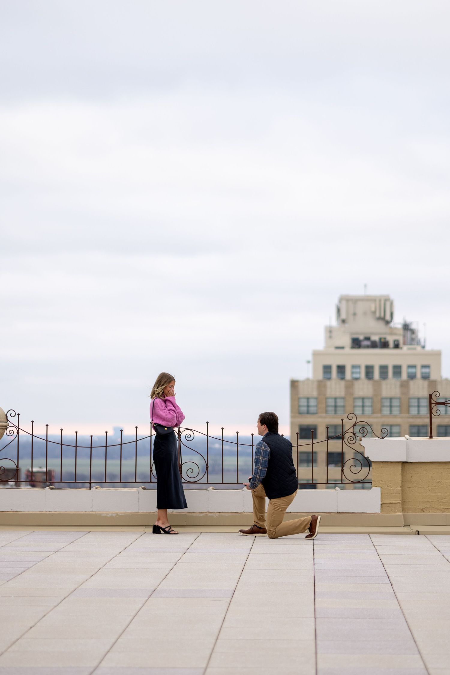Man kneeling in marriage proposal to woman in pink blouse on scenic urban rooftop terrace.