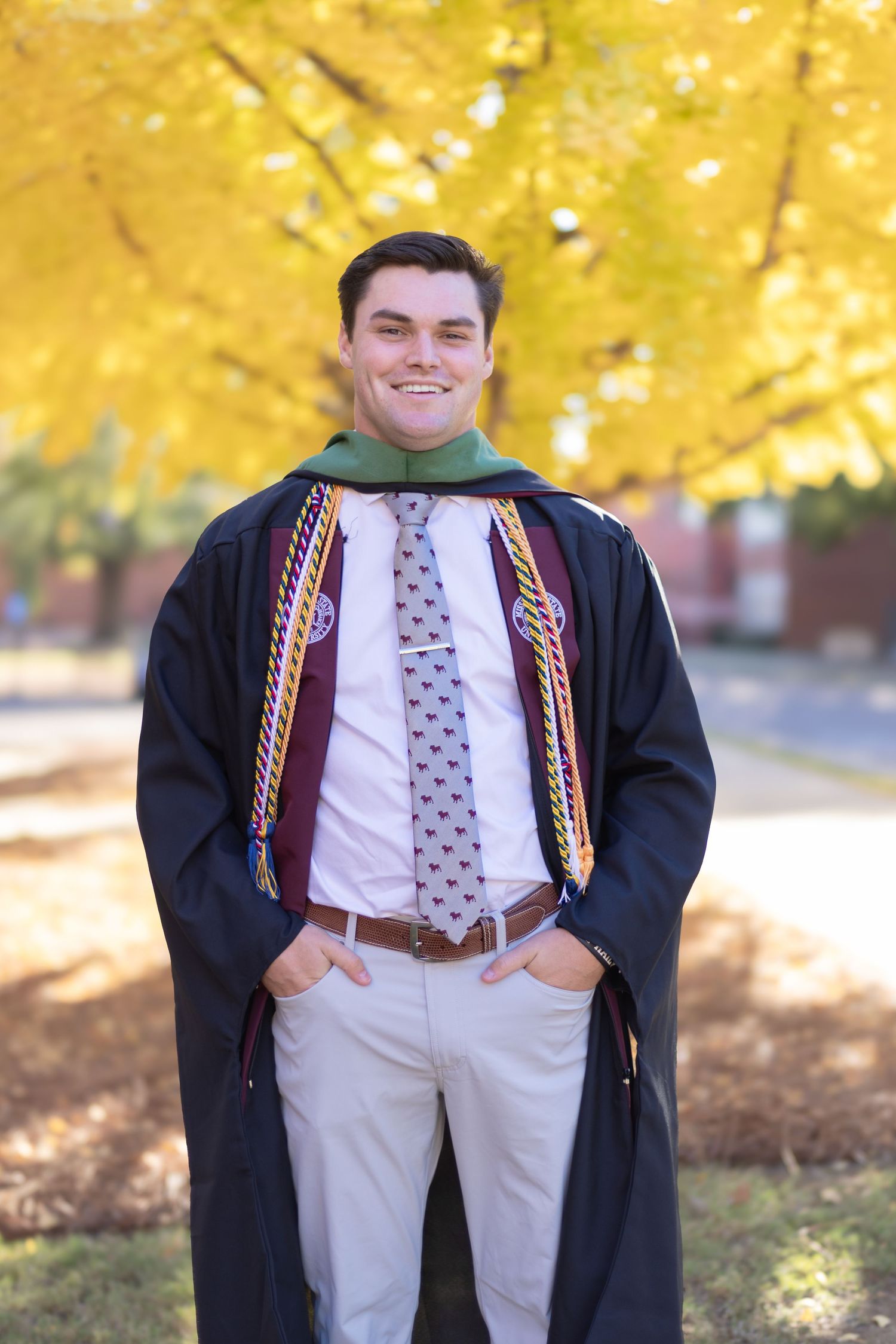 Graduate in black gown and colorful stoles poses against autumn foliage background.