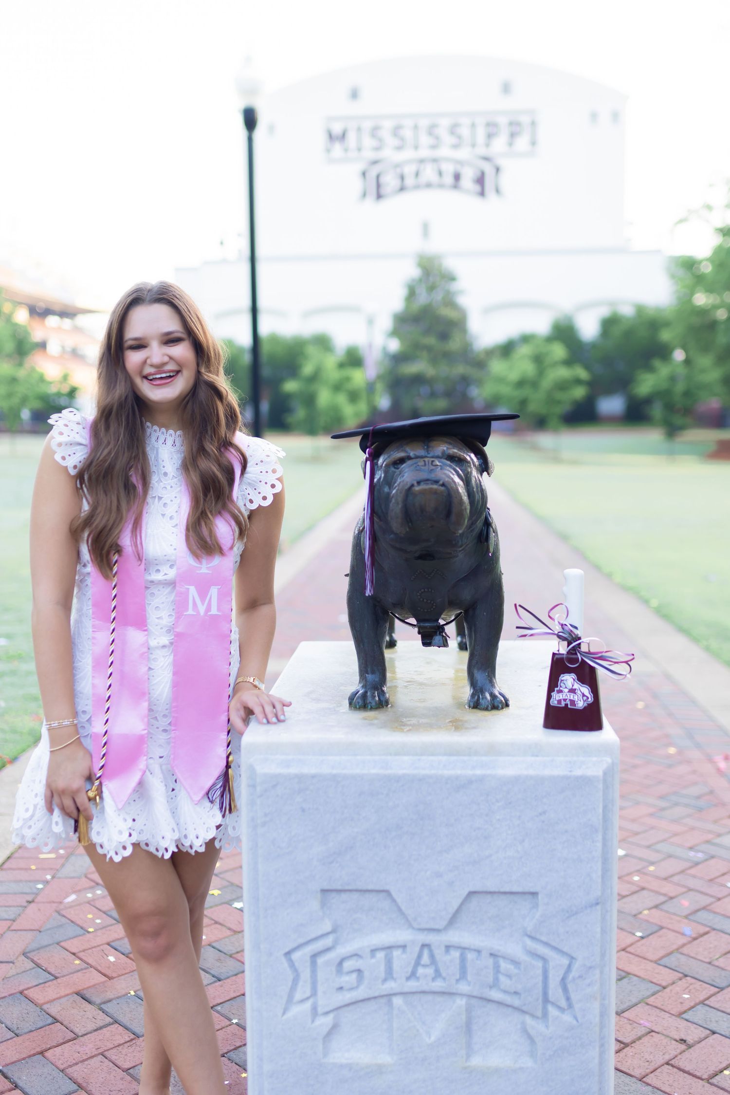 Graduate in pink outfit poses with Mississippi State bulldog statue on brick pathway.