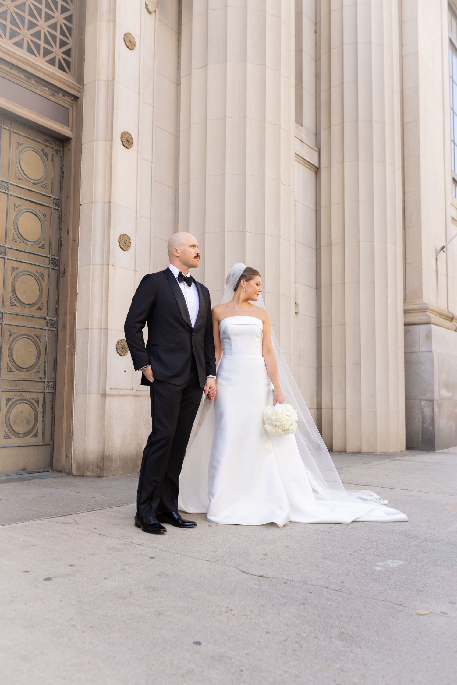 Bride and groom posing outside grand stone building, she in strapless white gown with veil, he in classic black tuxedo.