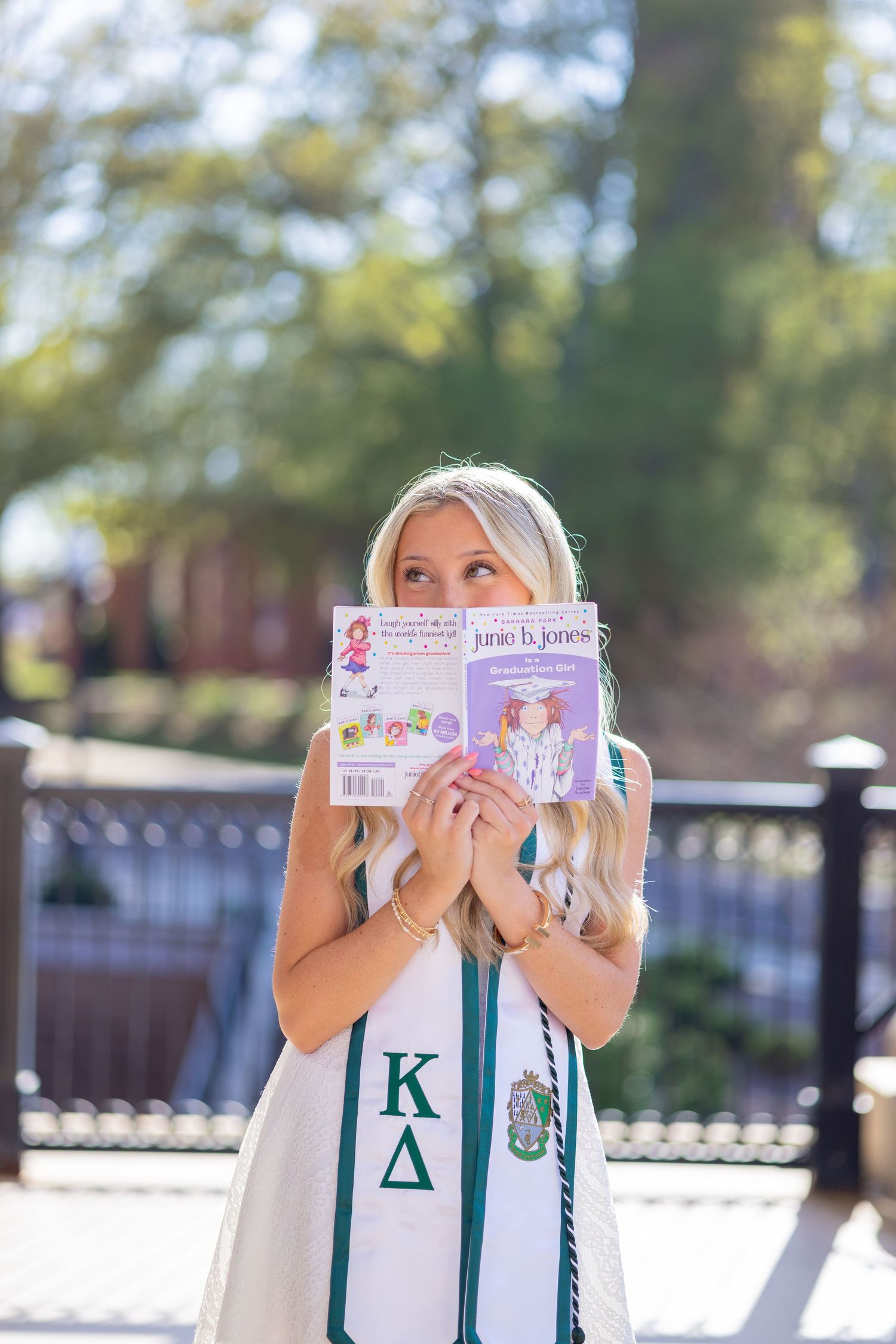 Blonde college graduate holding Junie B. Jones book, wearing Kappa Delta sash outdoors.