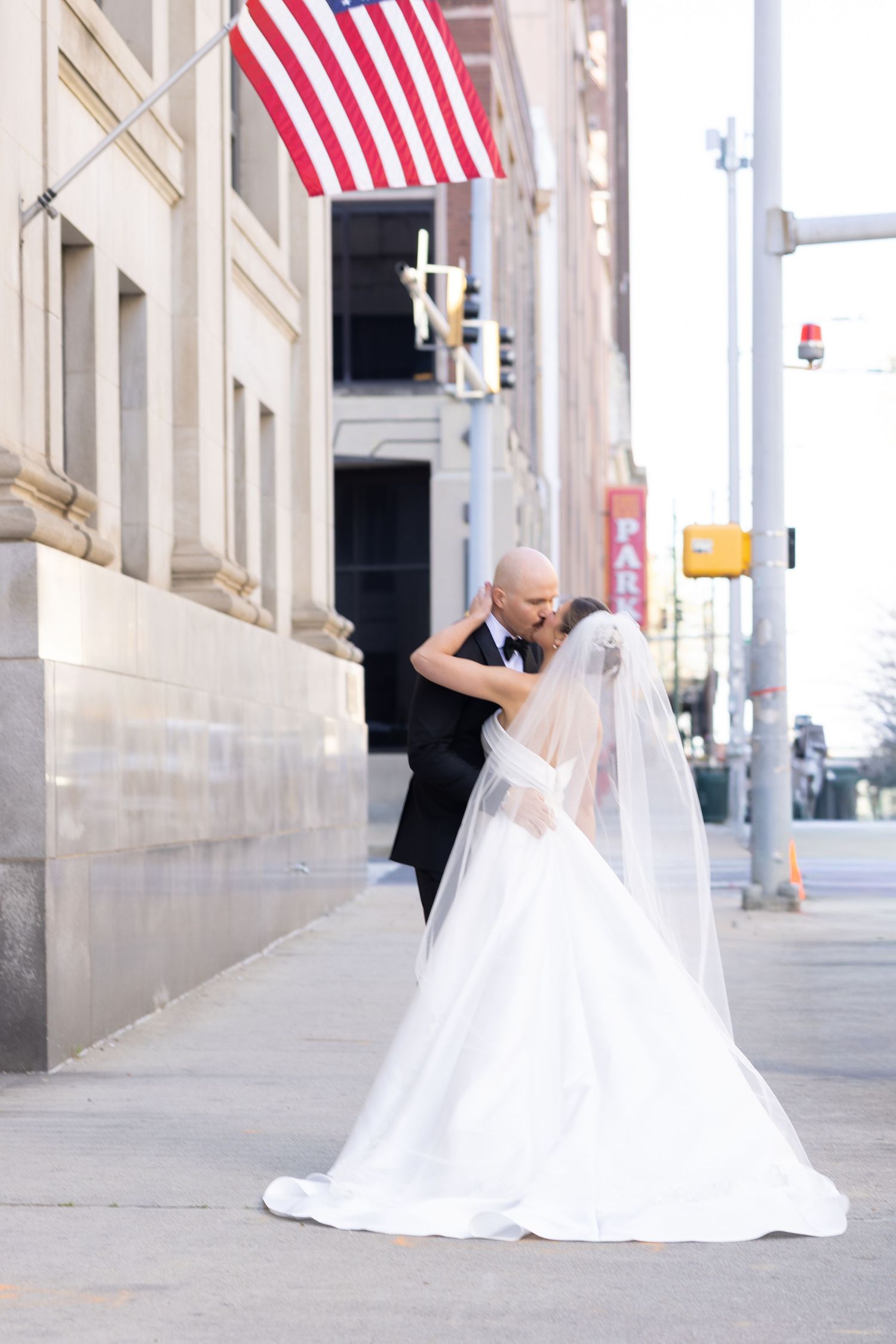 Bride and groom share a romantic kiss on a city sidewalk, bride in white gown with veil, groom in black tux, American flag above.