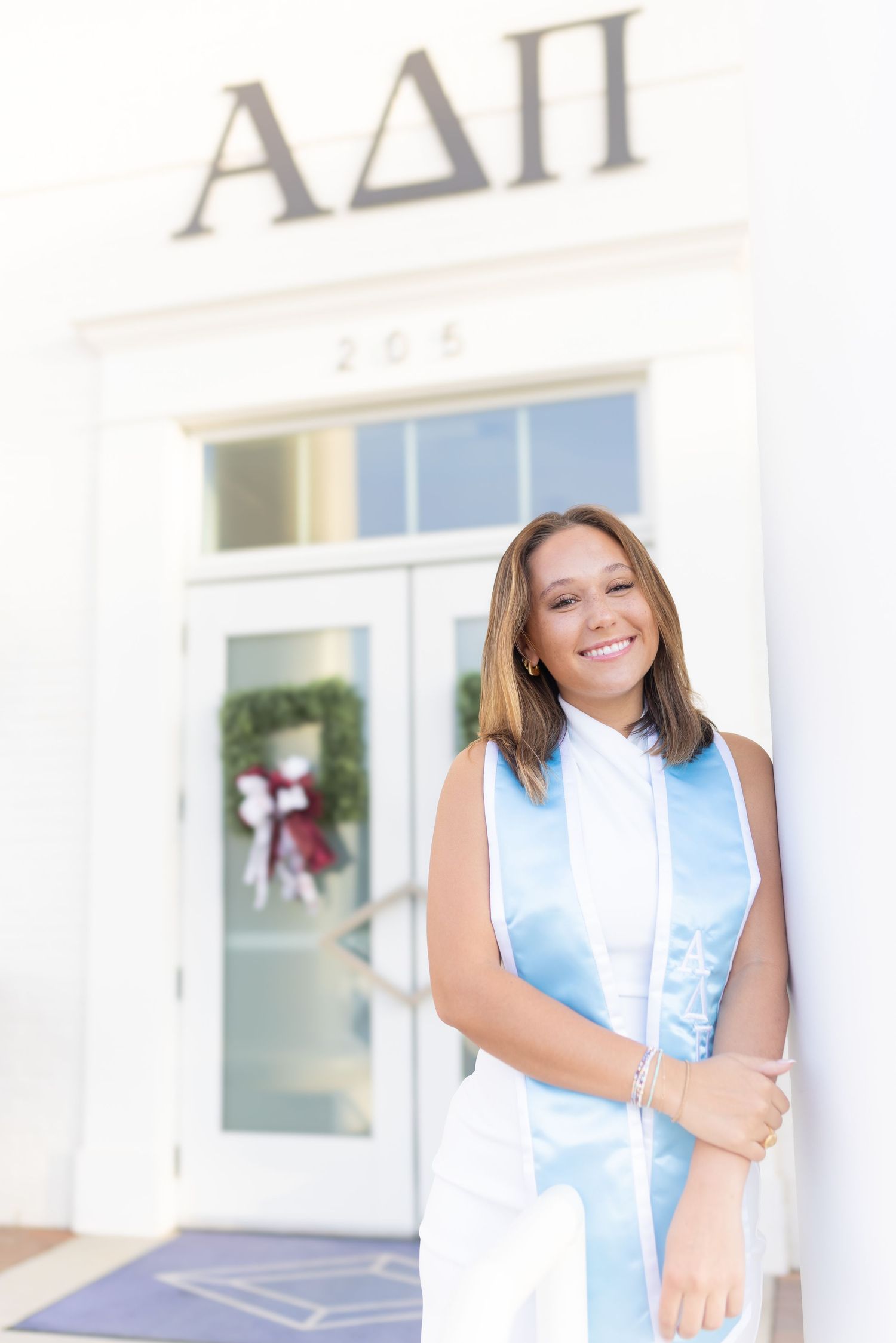 Woman in light blue sorority letters poses in front of white sorority house.