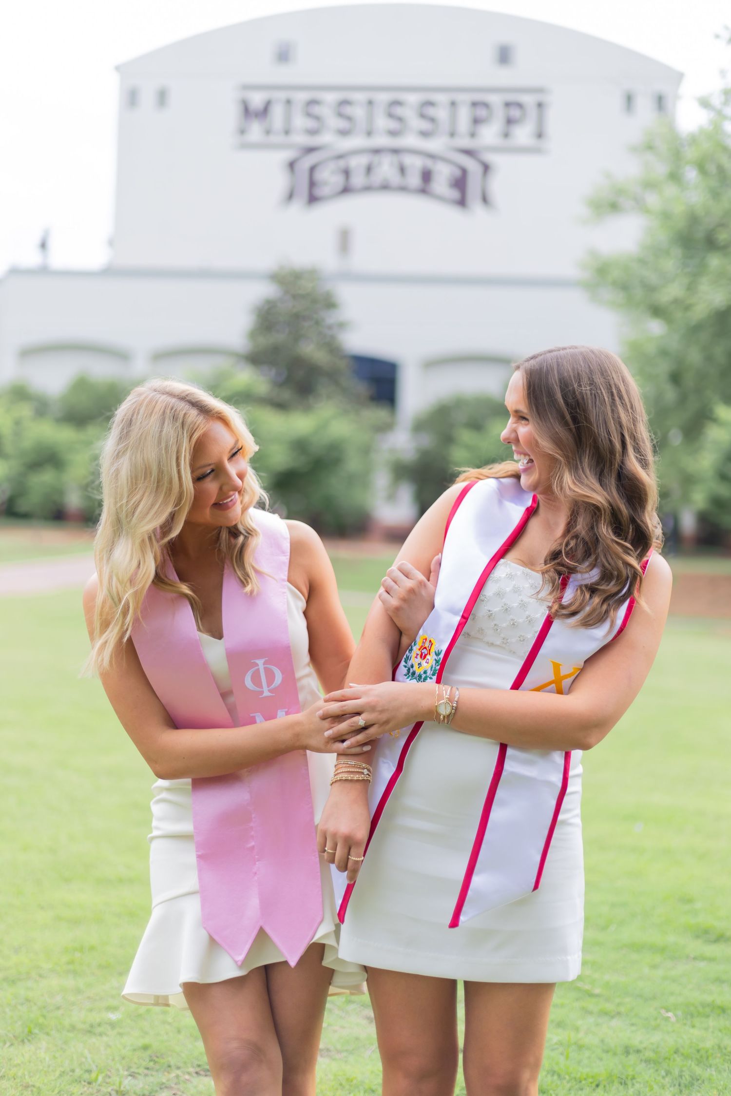 Two women in matching outfits embrace and laugh while posing on university grounds.