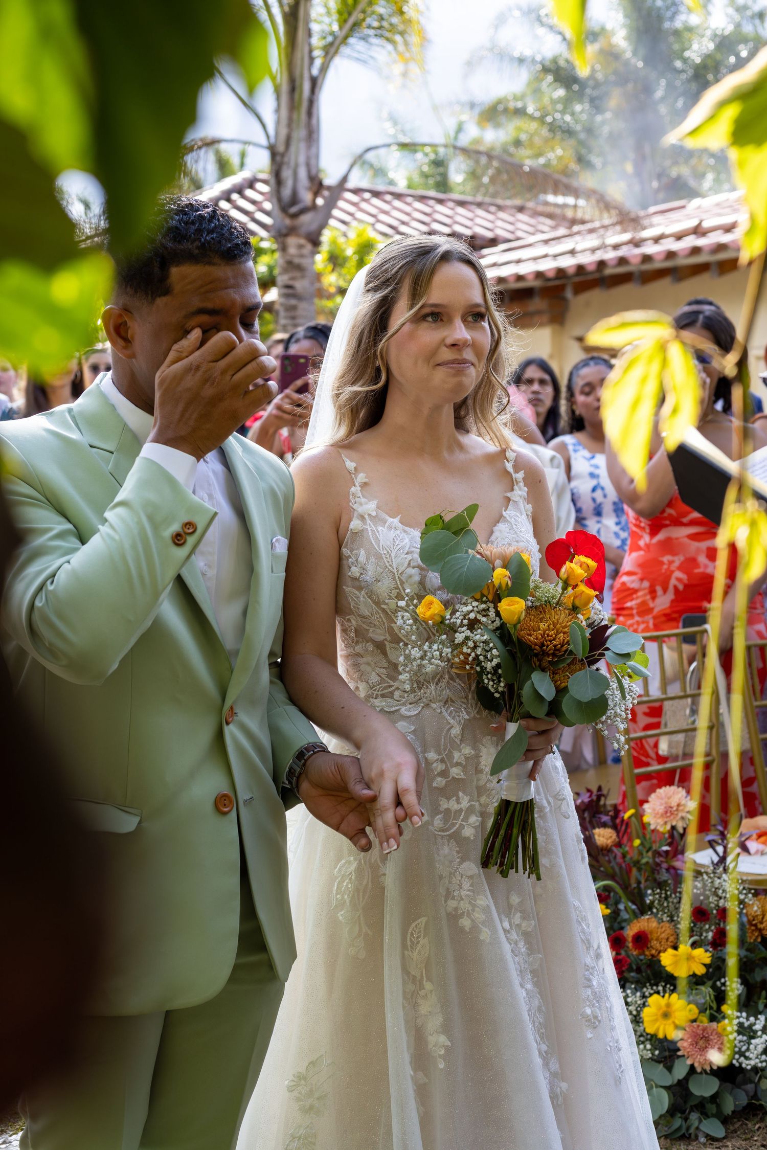 Emotional wedding moment captured in tropical garden setting with colorful floral arrangements.