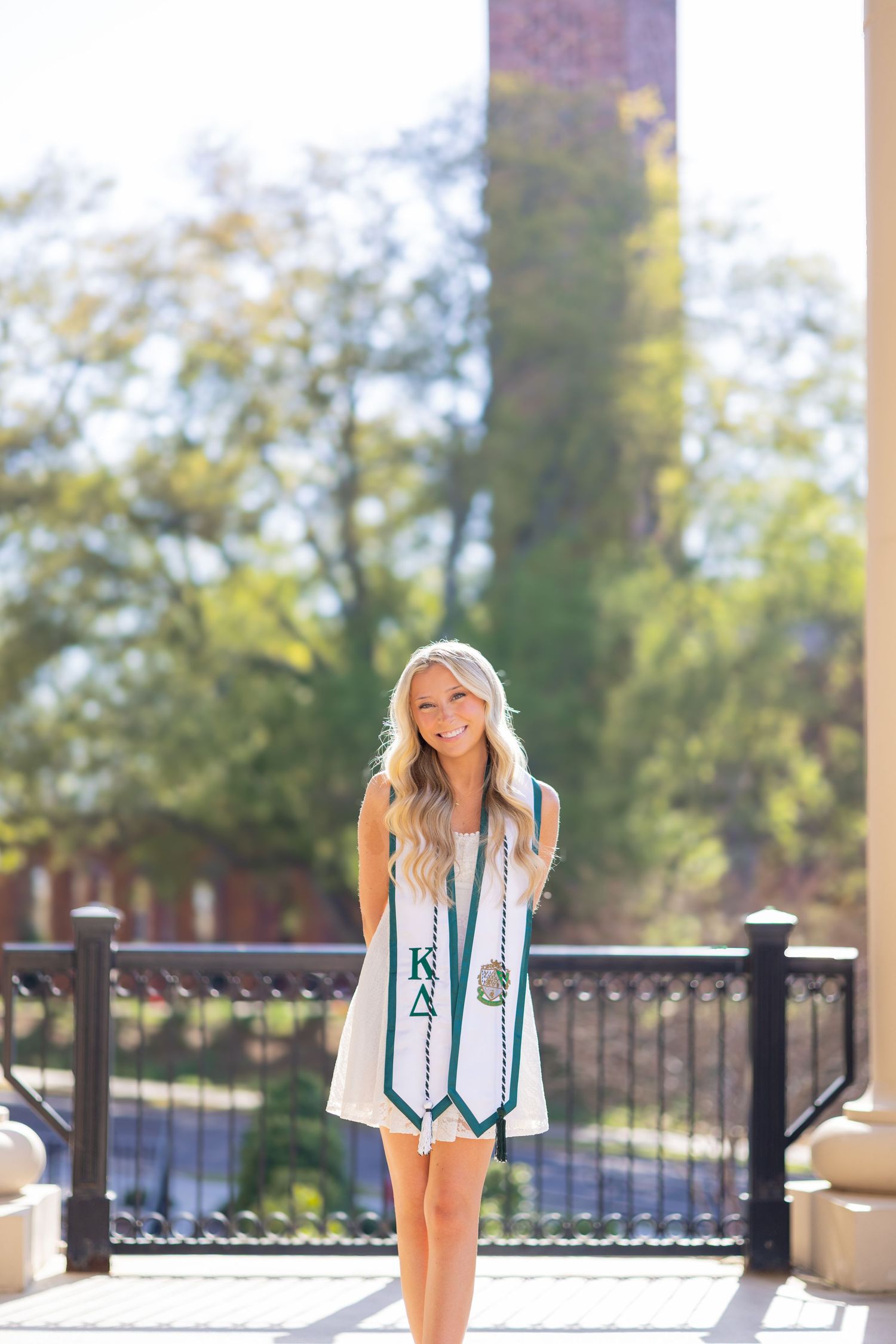 Smiling college graduate wearing Kappa Delta stole poses outdoors on campus in sunny spring weather.