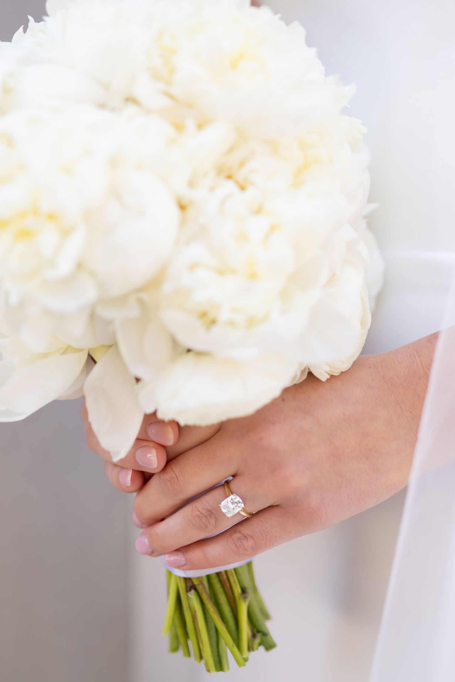 Bride holding white peony bouquet with elegant diamond engagement ring on her finger.