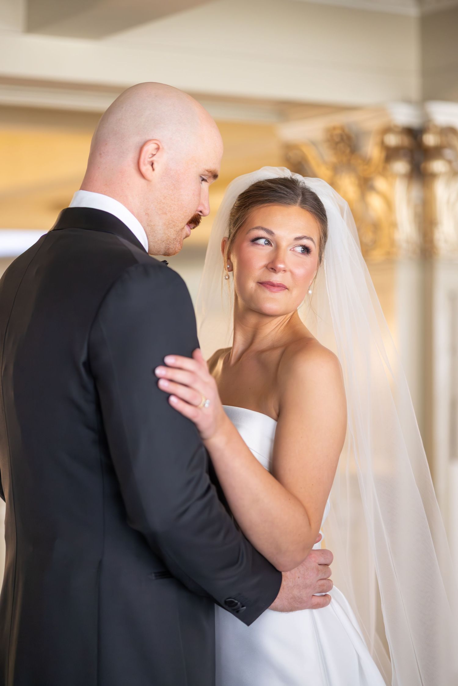 Bride in white gown and veil gazes at bald groom in black tuxedo during wedding dance.