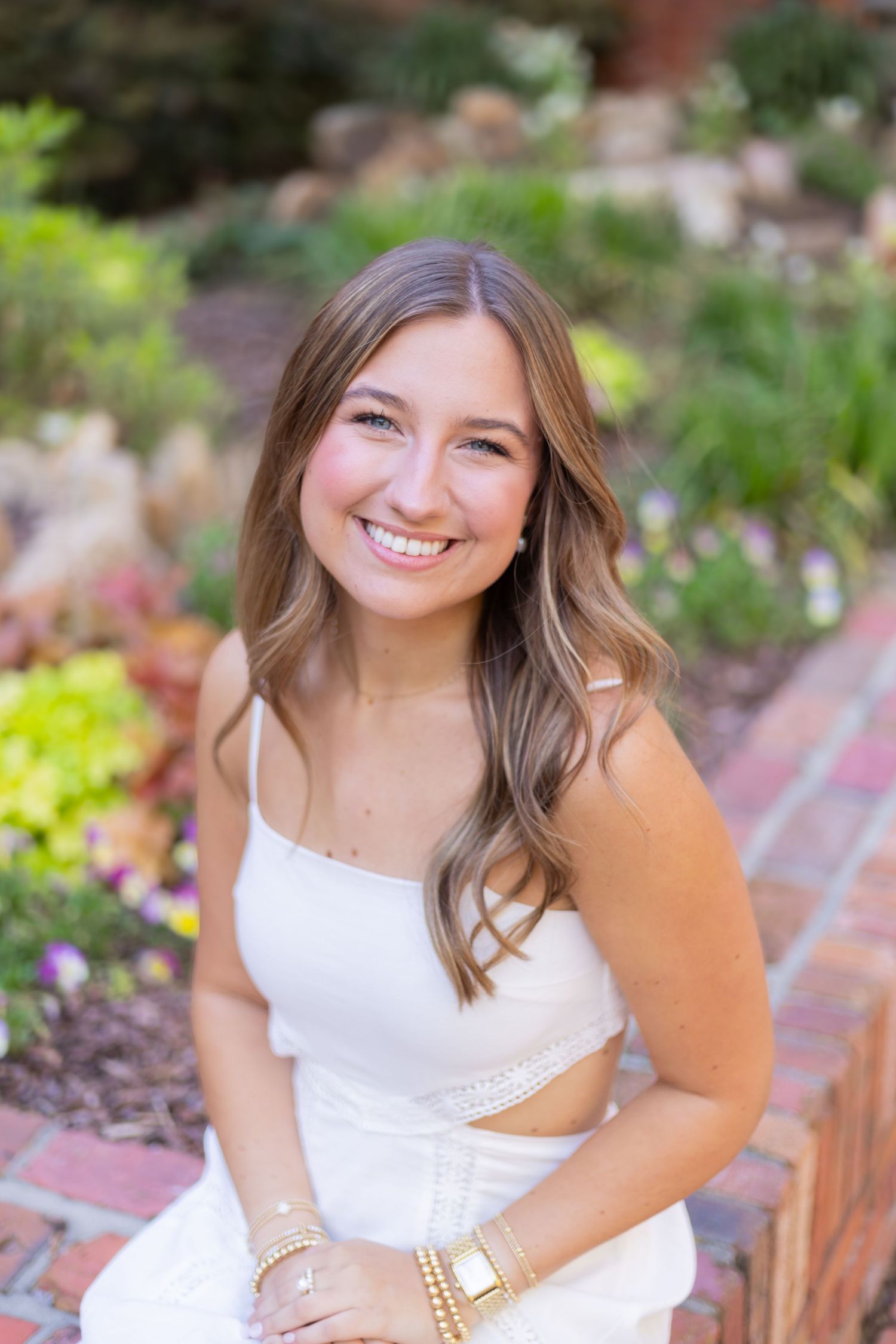 Graduate in white dress poses for portraits in a blooming garden setting.