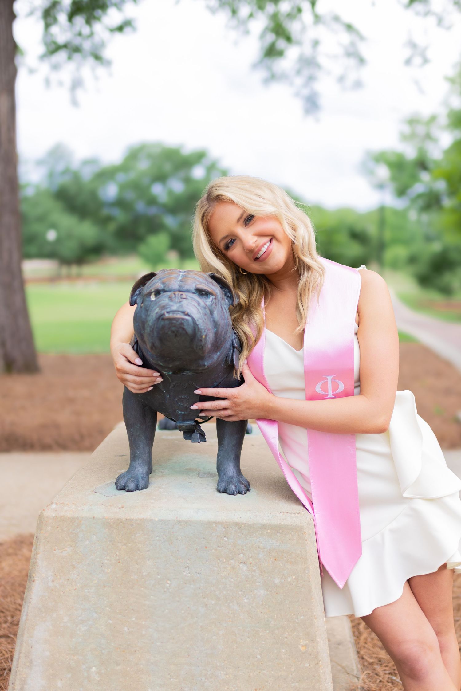 Graduate in white outfit interacts with bulldog statue during graduation photoshoot.
