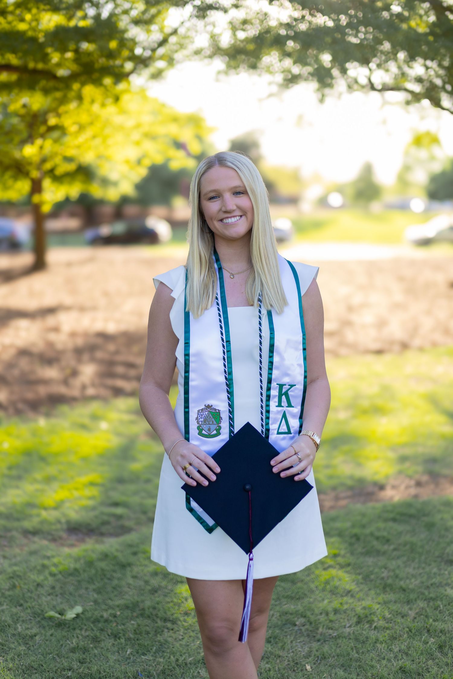 Graduate holds up decorated mortarboard cap with teal ribbons on grassy lawn during sunset.
