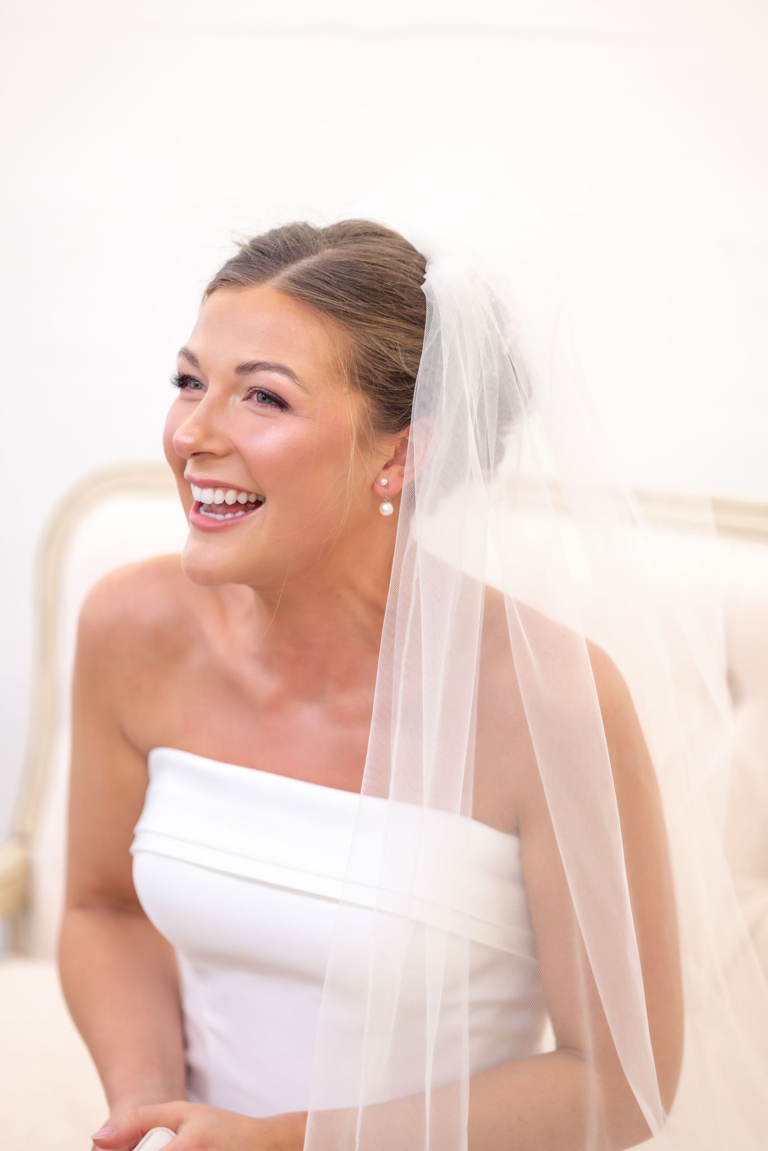 Smiling bride in white strapless gown and veil wearing pearl earrings on her wedding day.