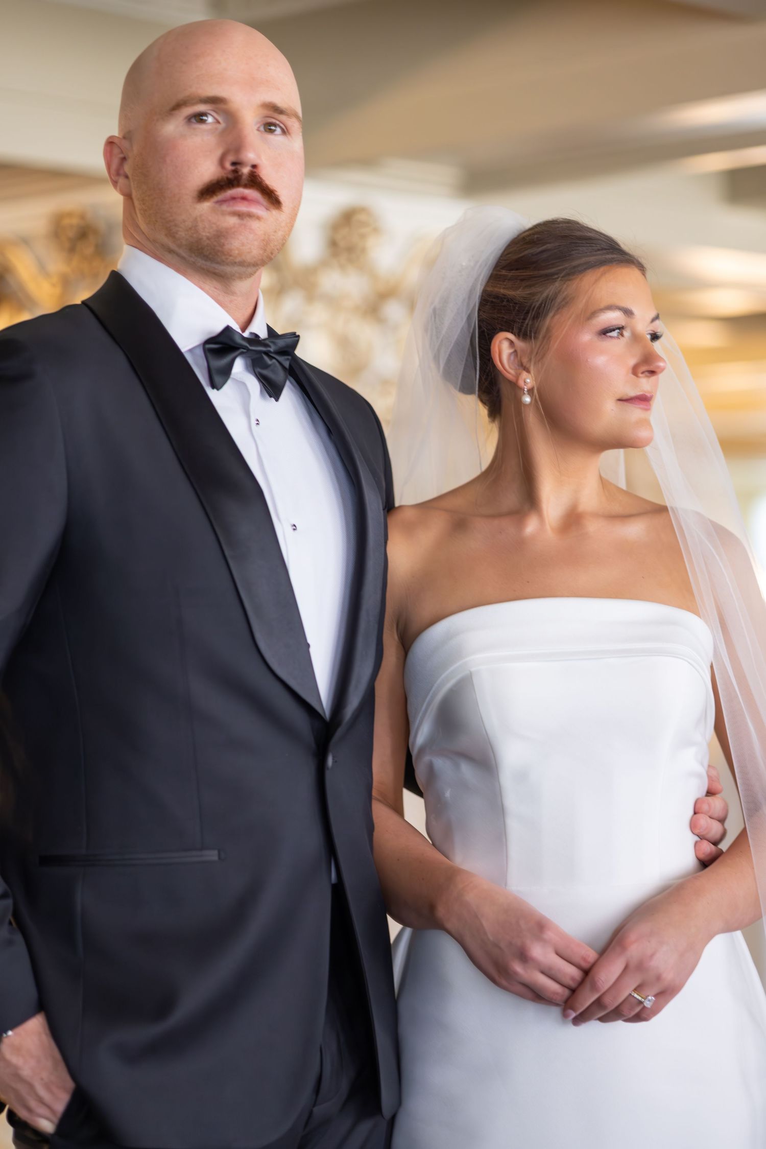 Bride in white strapless gown and veil stands beside bald groom in black tuxedo with bow tie at wedding ceremony.