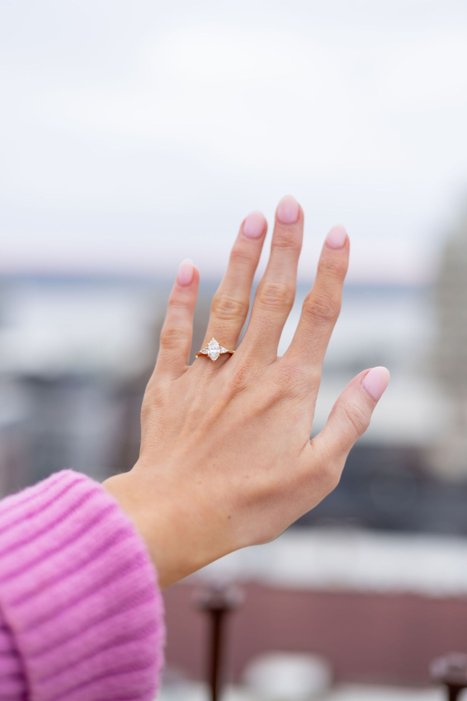 Close-up of a hand with pink nails wearing a rose gold diamond engagement ring, pink sweater sleeve visible.