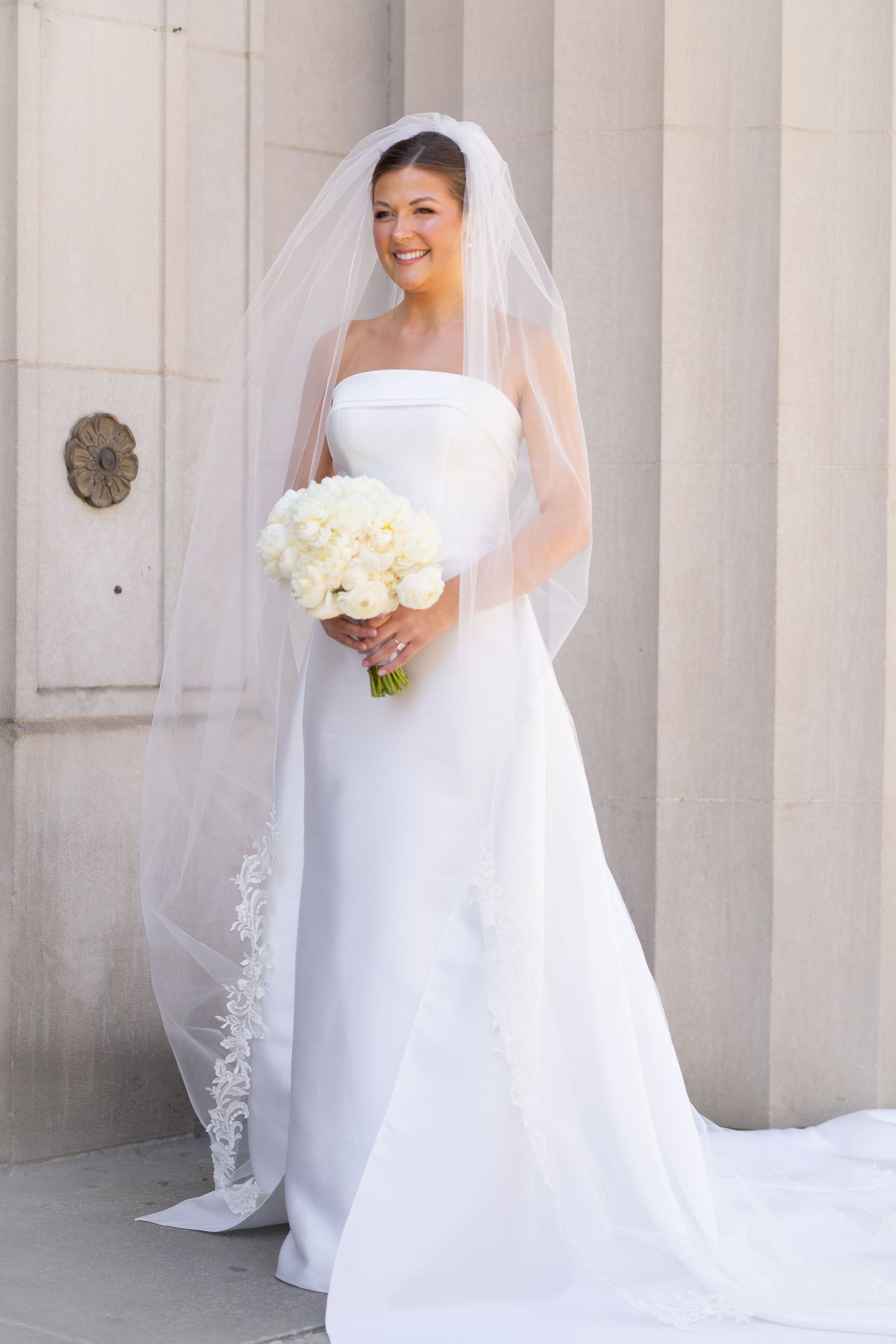 Smiling bride in white strapless gown and lace-trimmed veil holds white peony bouquet beside marble columns.