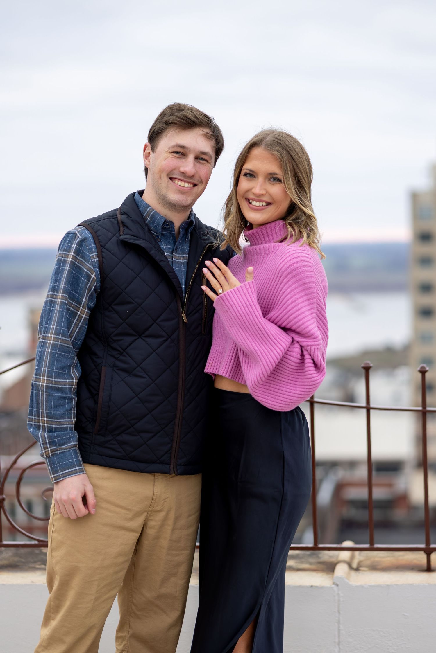Happy couple posing on a rooftop, woman in pink sweater and man in navy vest, with a cityscape backdrop.