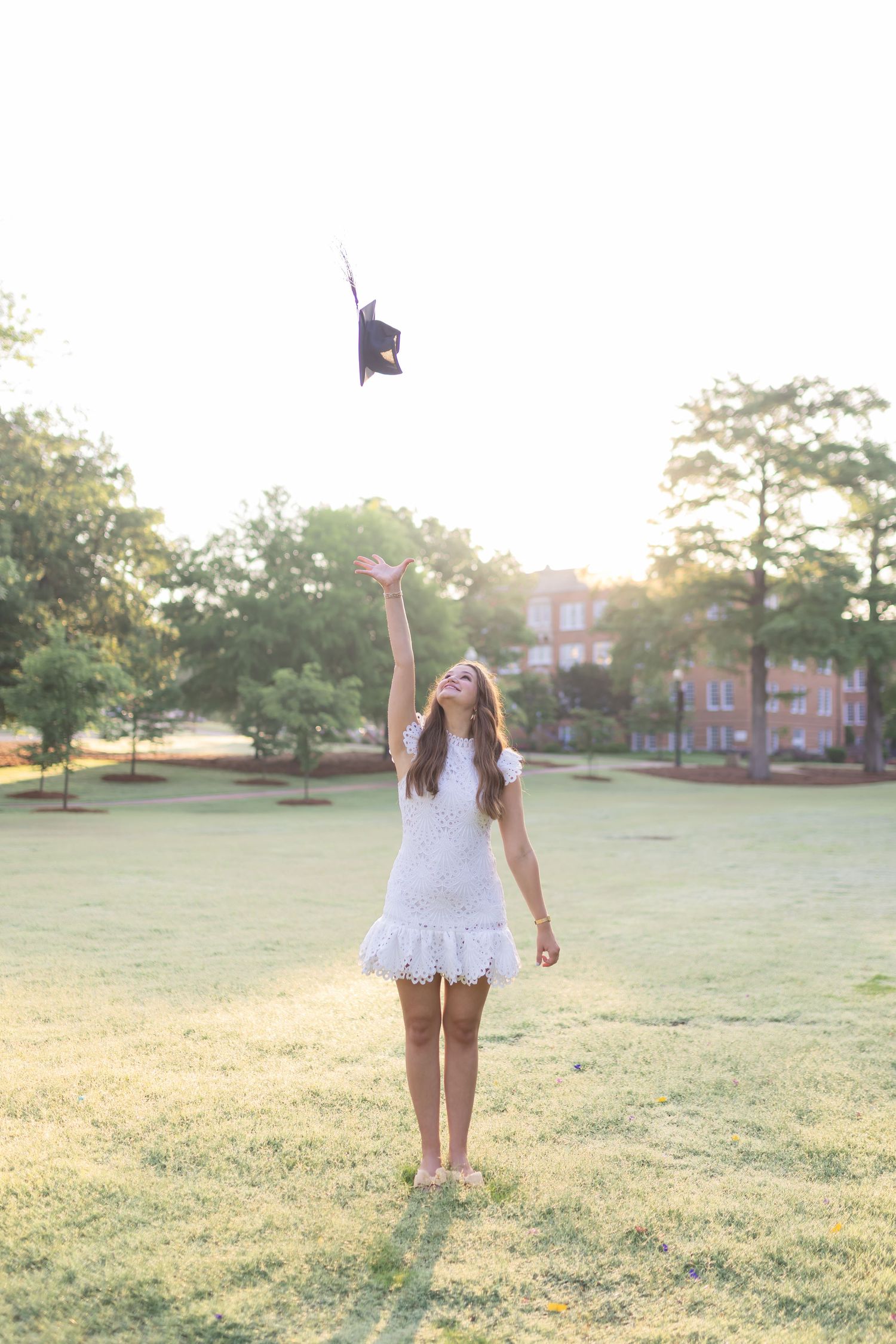 Graduate in white dress celebrates with cap toss in sunny field at sunset.