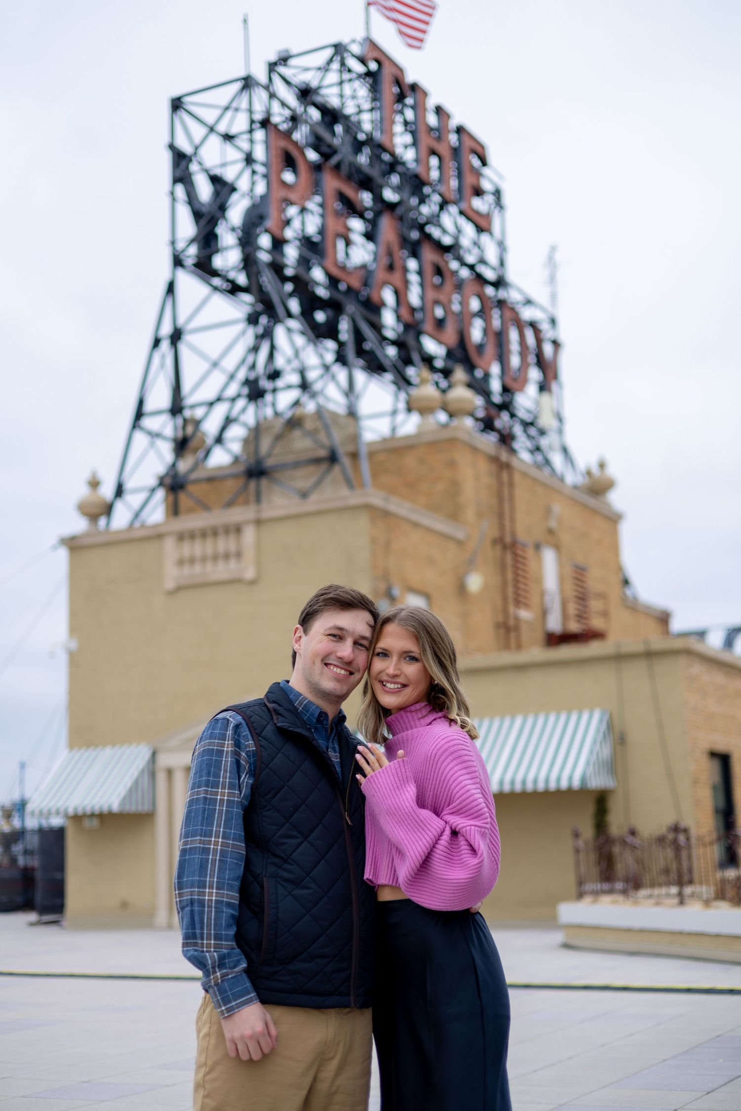 Couple smiling together in front of The Peabody Hotel sign outdoors, woman wearing pink sweater, man in navy vest.