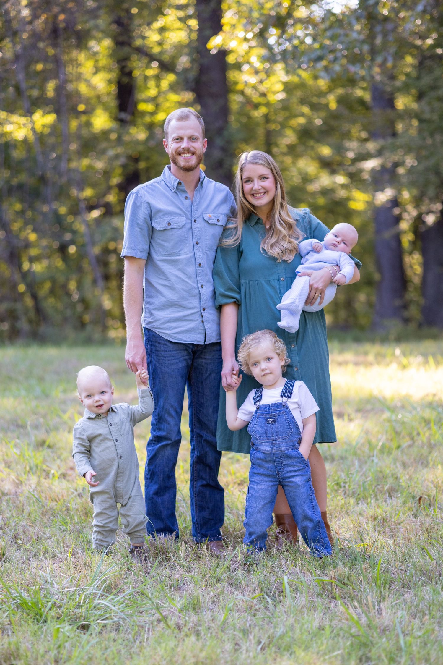 Family poses together in a grassy field during sunset with small children in matching outfits.