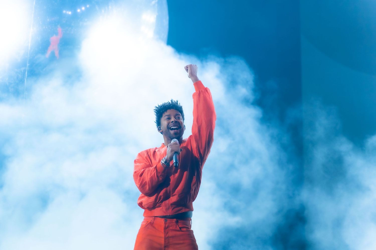 Person in red jacket stands triumphantly against dramatic cloudy blue sky background.