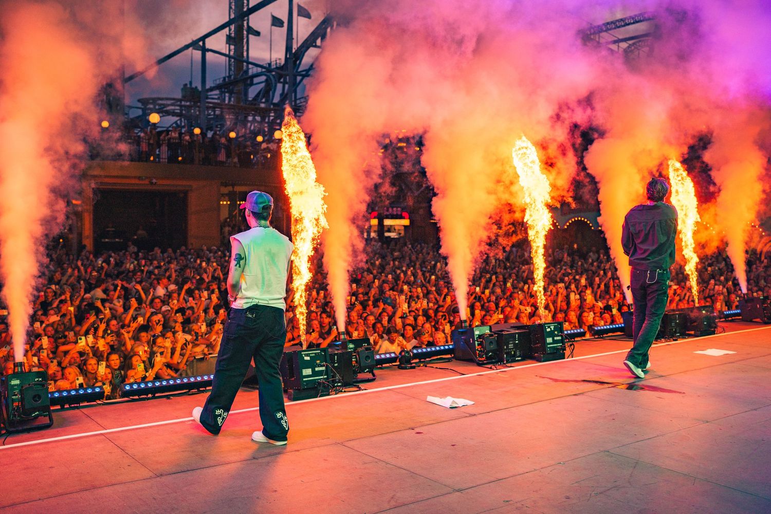 Dramatic concert stage with bright orange and pink pyrotechnics illuminating performers and crowd during live performance.