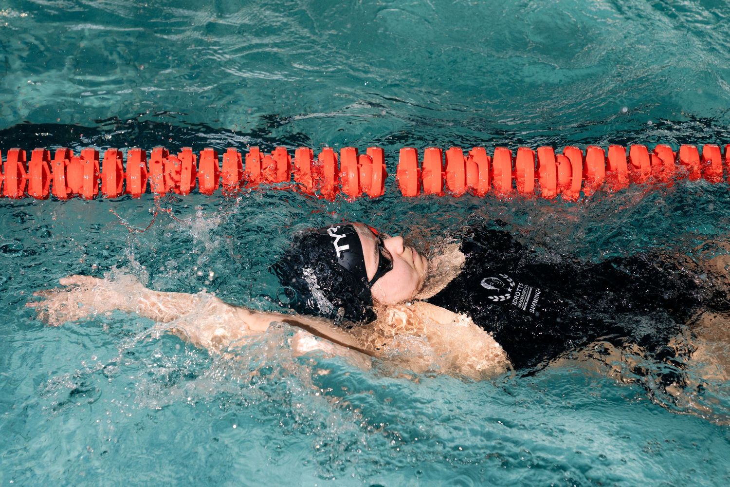 Competitive swimmer in black swimsuit and goggles performing backstroke in a pool with orange lane dividers.