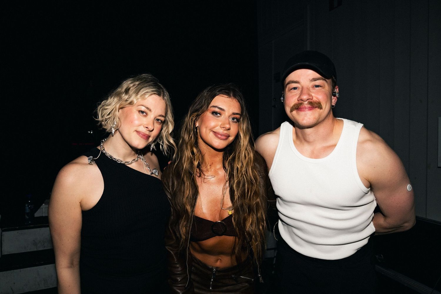 Three young adults smiling together in a dark venue, two women and one man posing for a photo.