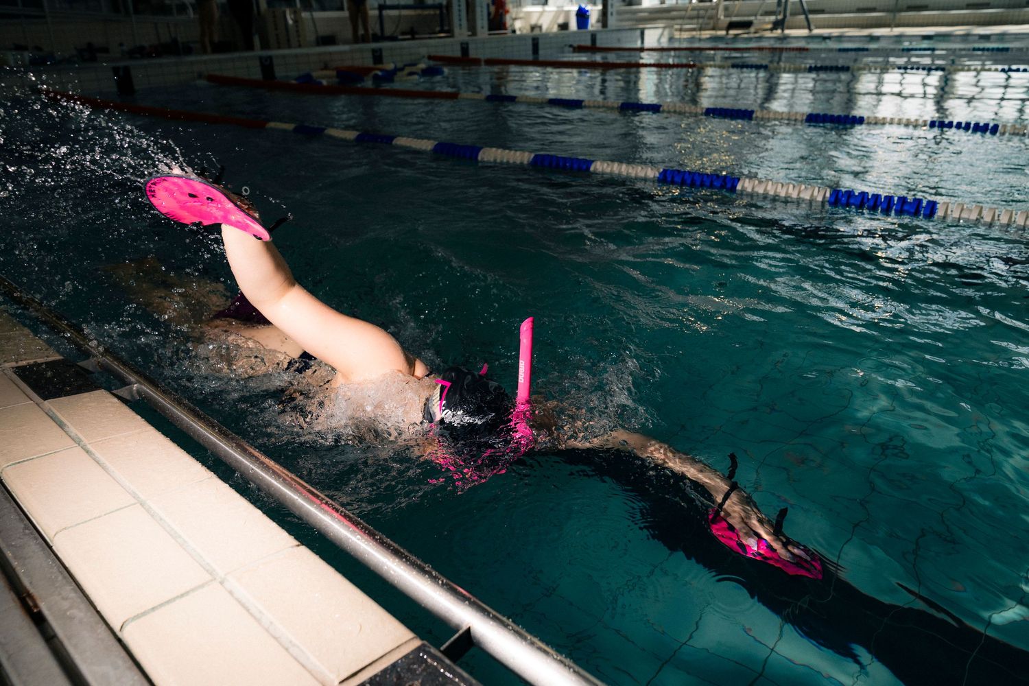 Swimmer diving into an indoor pool lane with pink swim cap and fins, creating splashes in a competitive swimming facility.