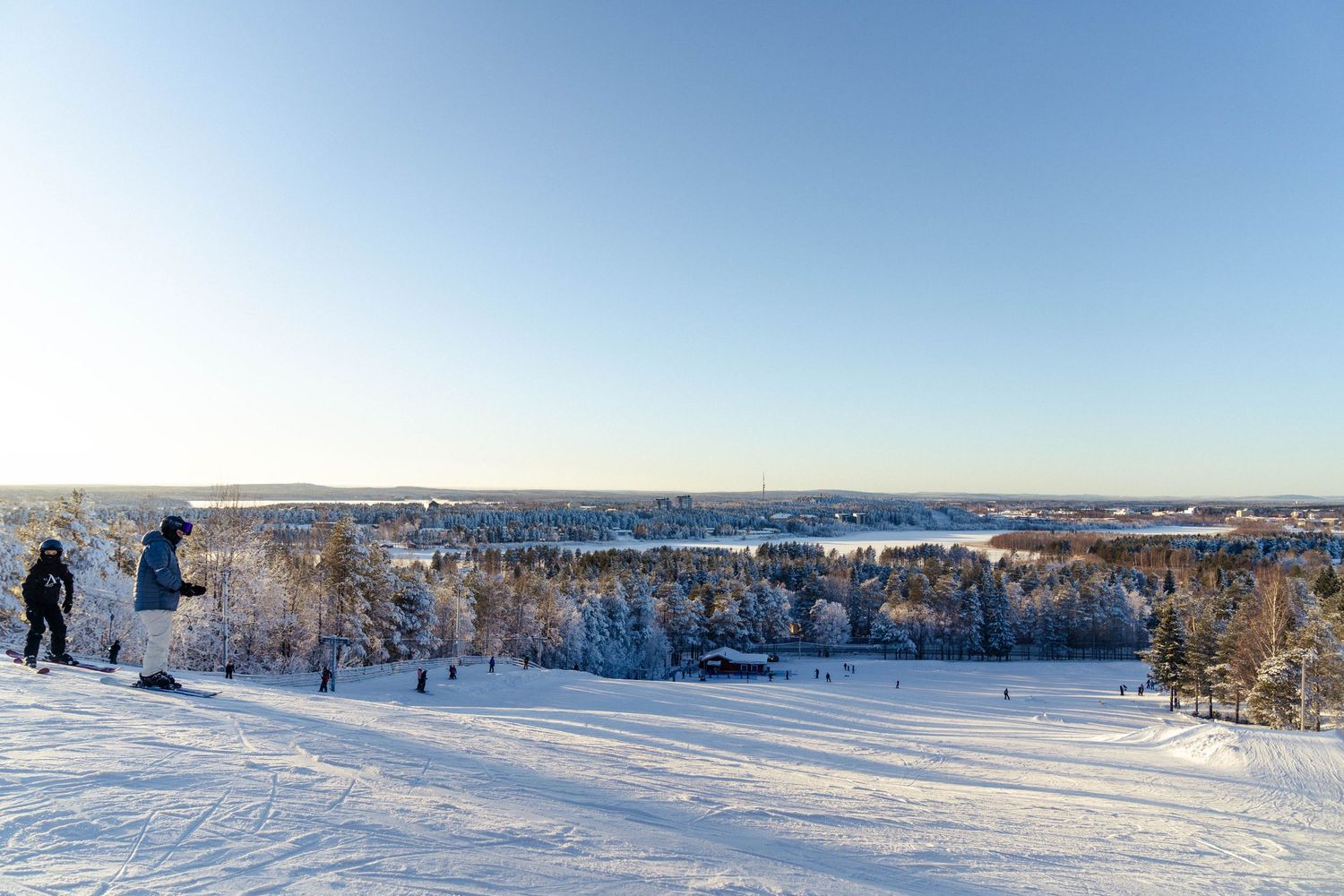 A panoramic winter landscape showing people ice fishing on a frozen lake surrounded by snow-covered trees and shoreline.