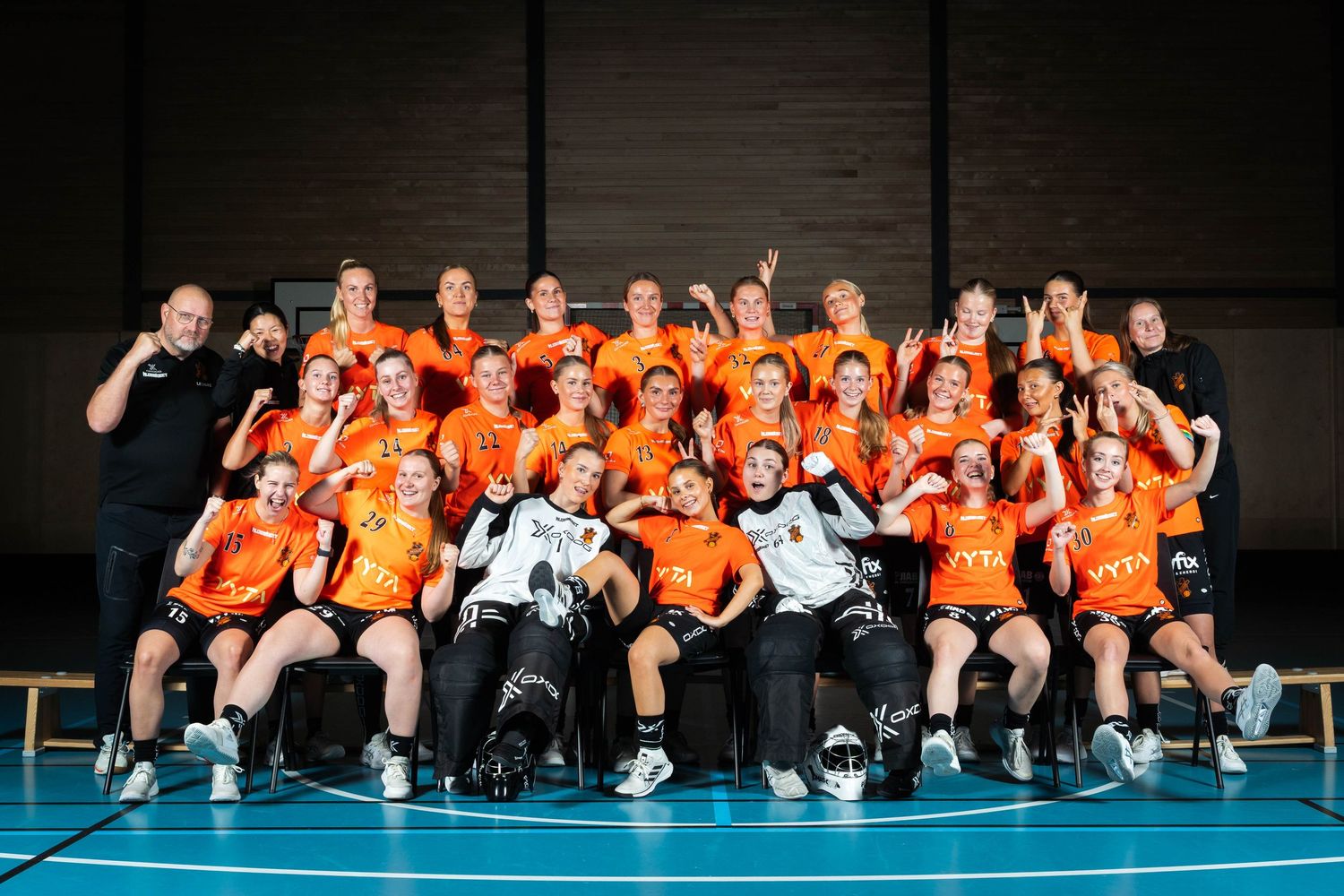 Sports team in orange jerseys poses together on indoor court floor for group photo.