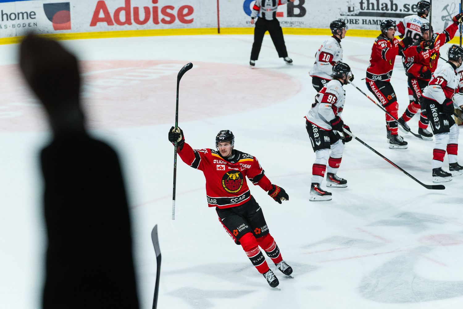 Hockey players in red jerseys skate on ice during a competitive game with spectators watching from the sidelines.