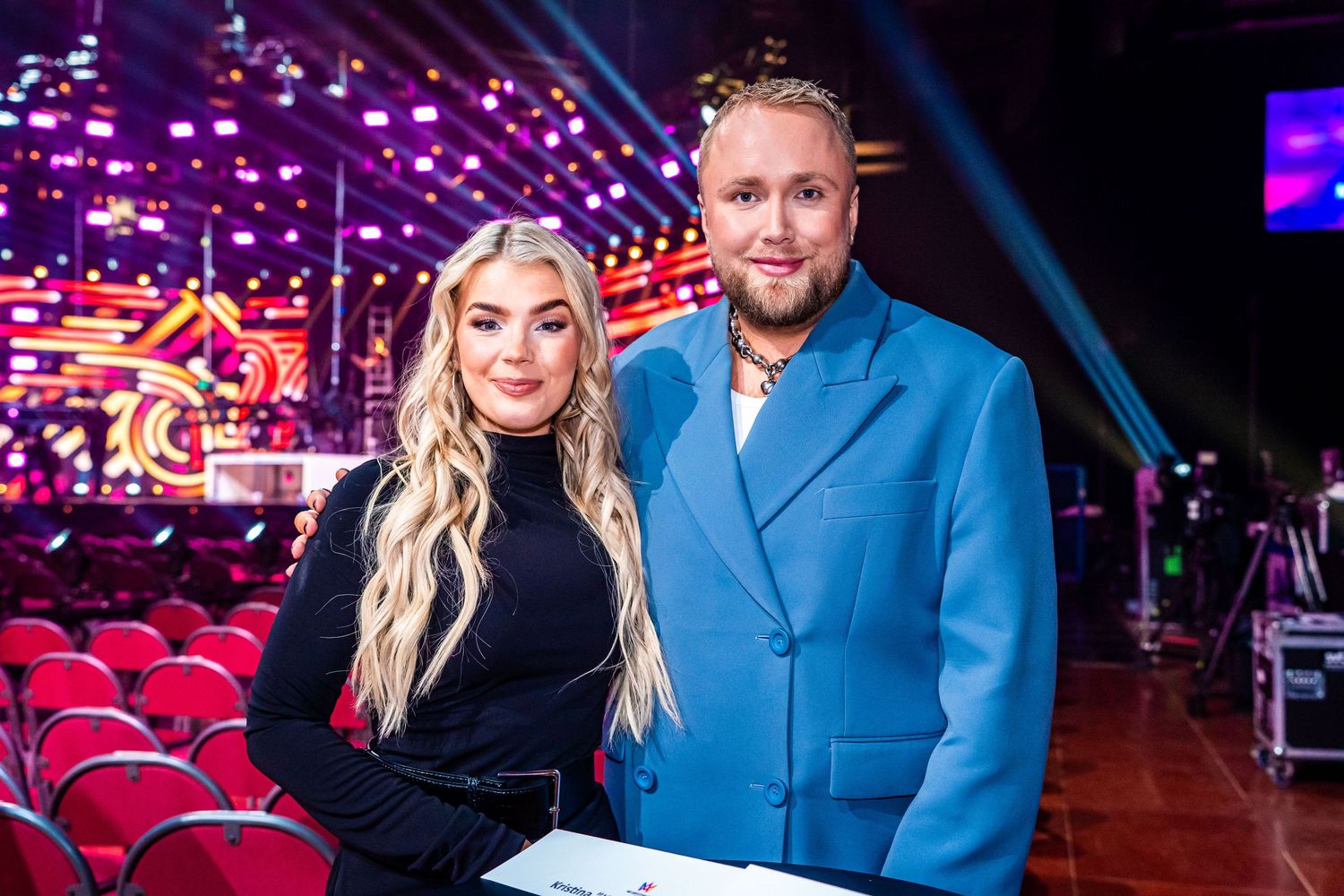 Two people pose together in front of a stage with red seats and purple lighting effects in the background.