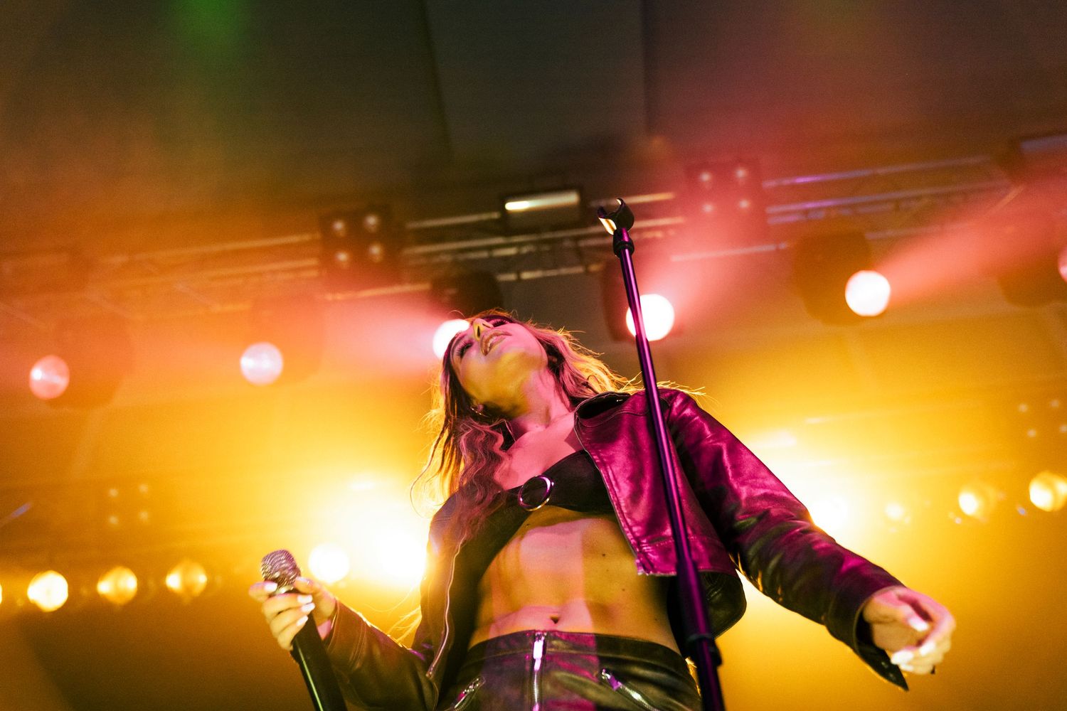 Female singer performing on stage under golden lights, wearing a cropped jacket, passionately singing into a microphone.