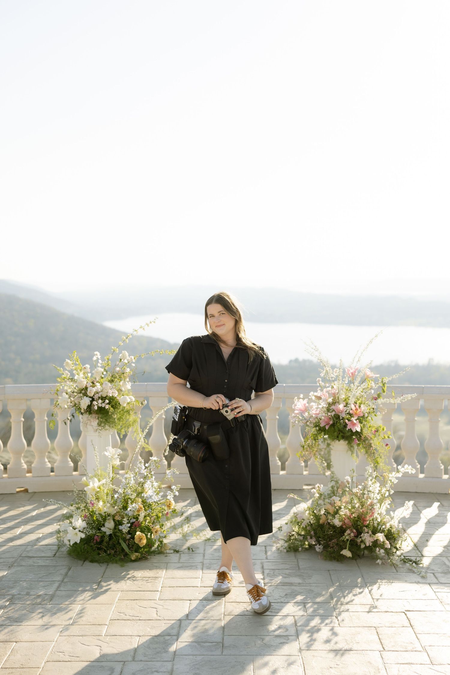Woman in black dress holding camera on stone terrace with floral arrangements and scenic lake view.
