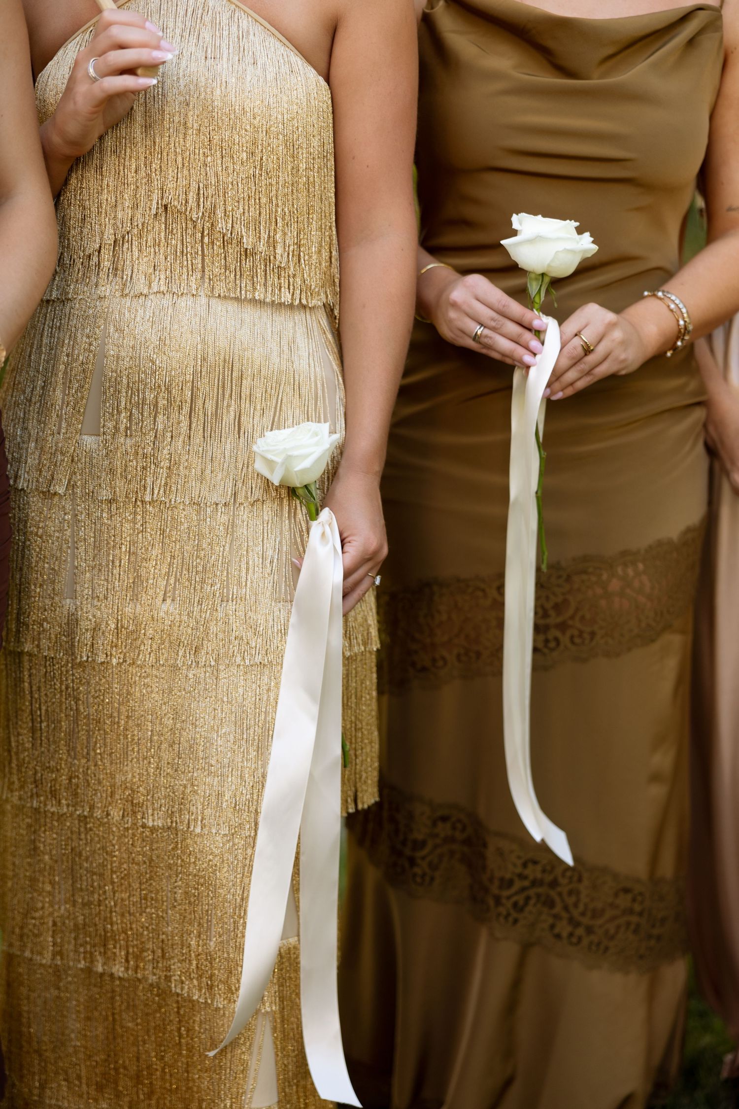 Two elegant dresses, one gold with fringe and one brown satin, shown with white roses and ribbons at a formal event.