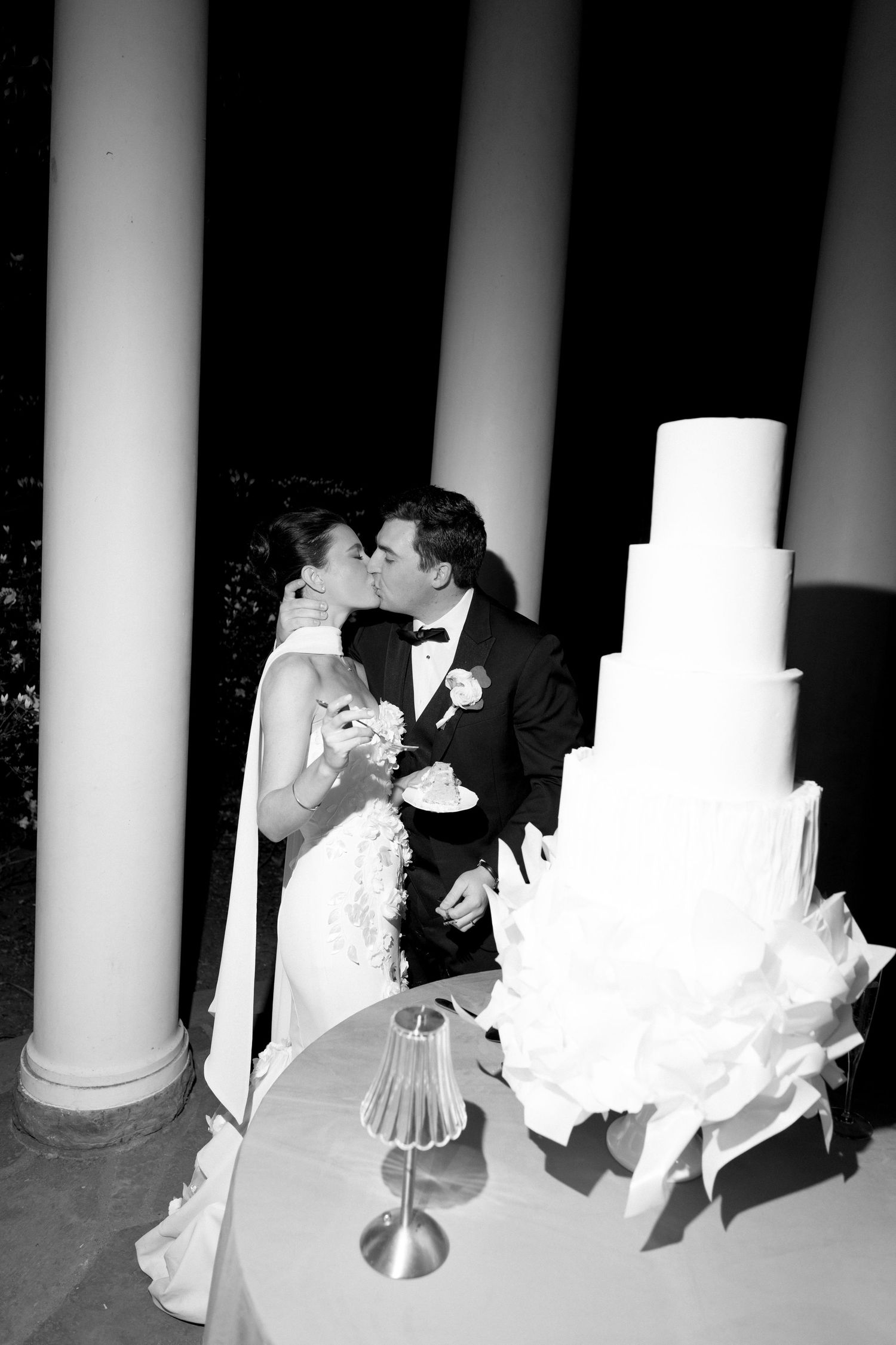 Black and white photo of a bride and groom kissing beside a tall elegant tiered wedding cake at their reception.