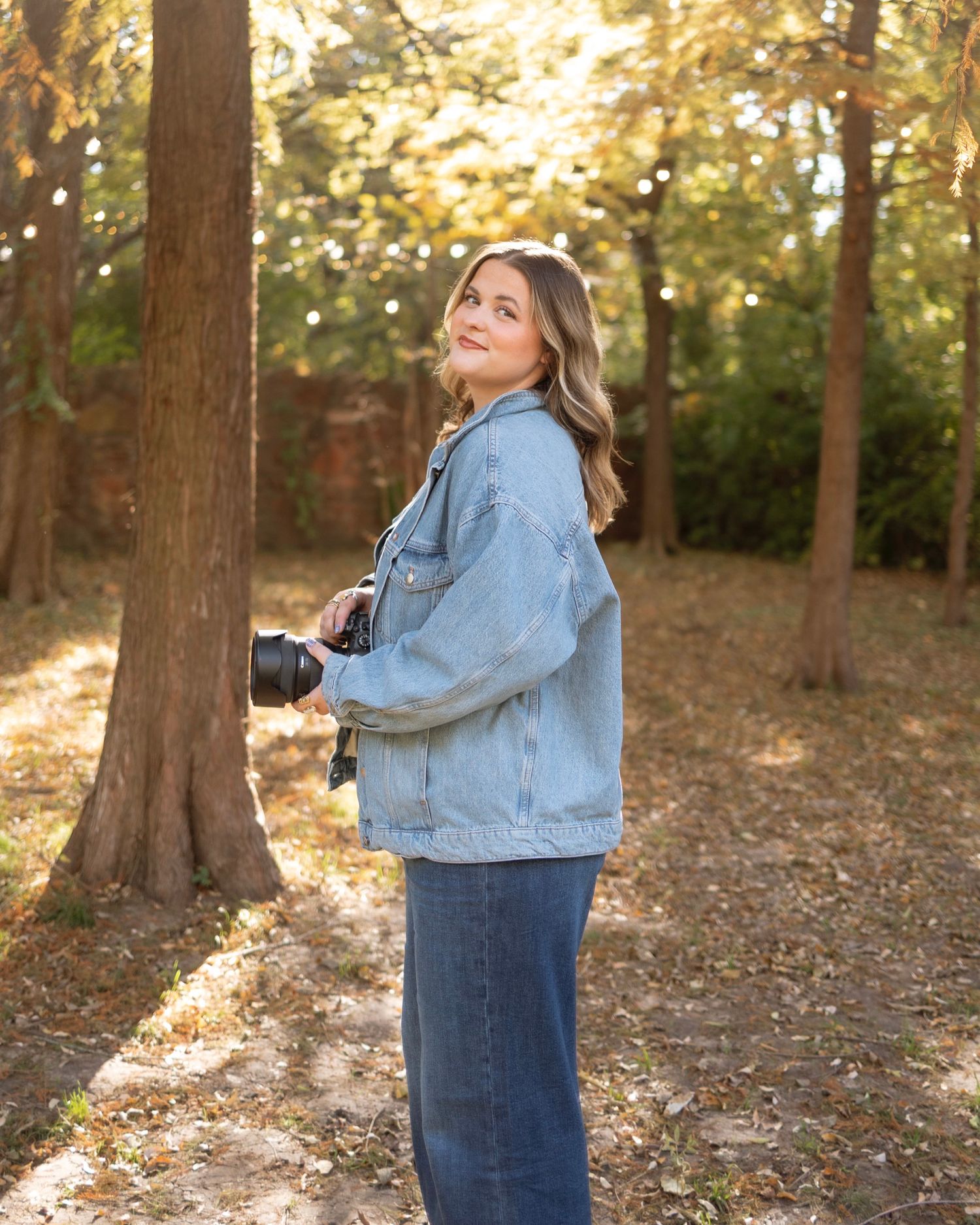 A photographer in denim clothing stands in a wooded area during golden hour capturing photos among fallen leaves.
