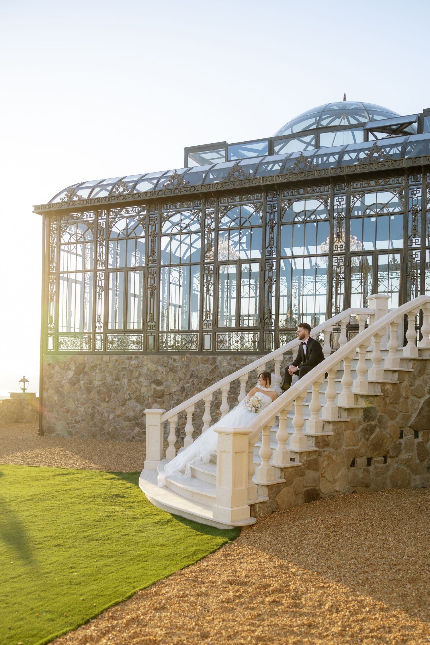 Bride and groom sit on white balustrade stairs beside an ornate Victorian glass greenhouse at golden hour.