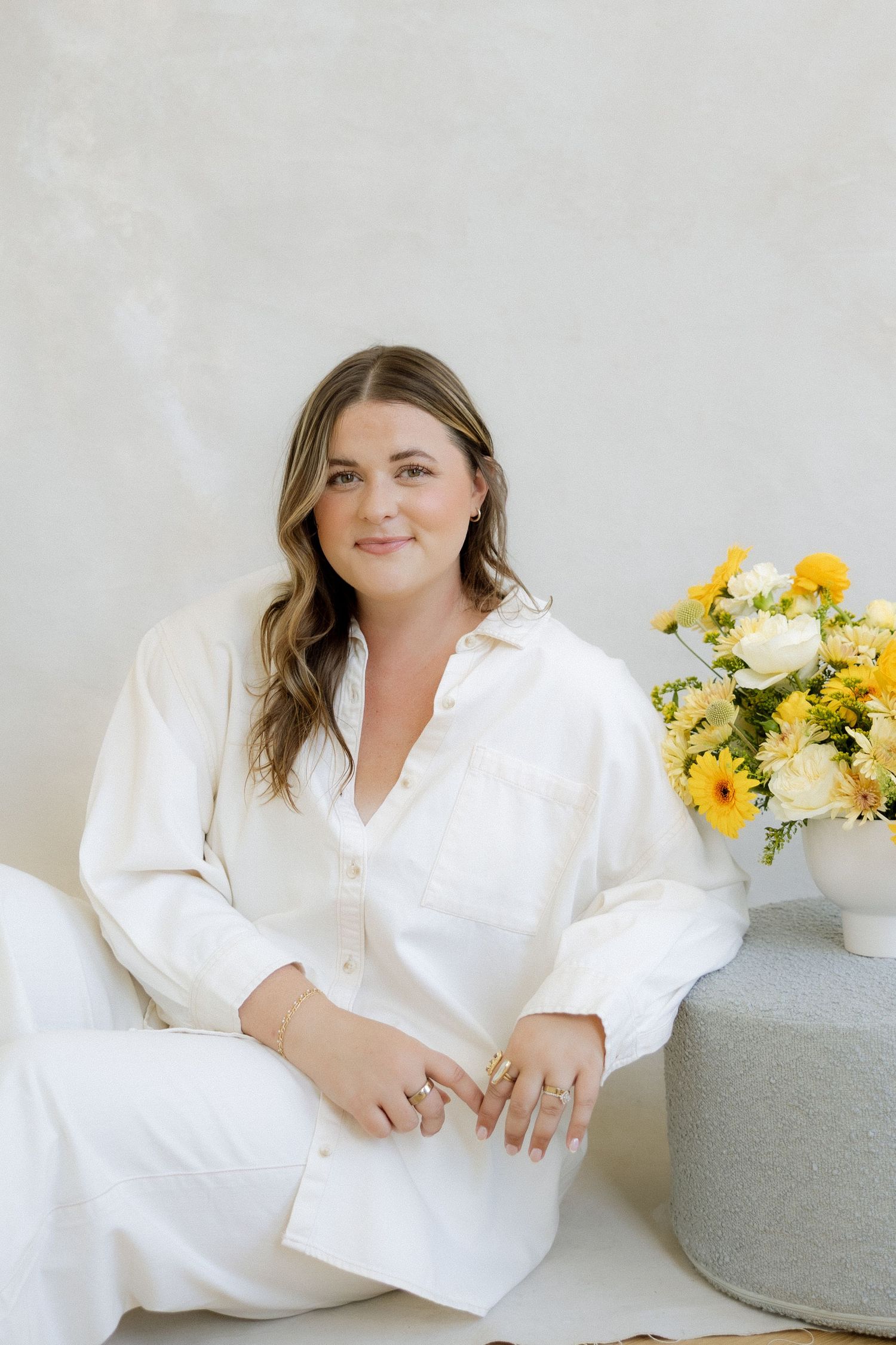 Woman in white linen outfit sitting beside yellow and white flower arrangement on a neutral background.