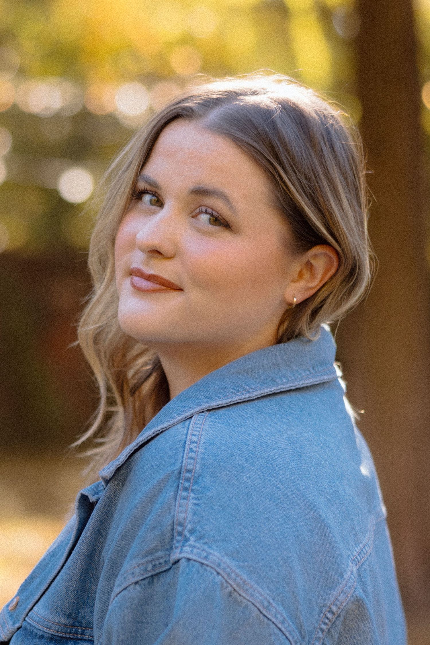 A woman in a denim jacket poses outdoors with a playful expression against a sunlit natural background.
