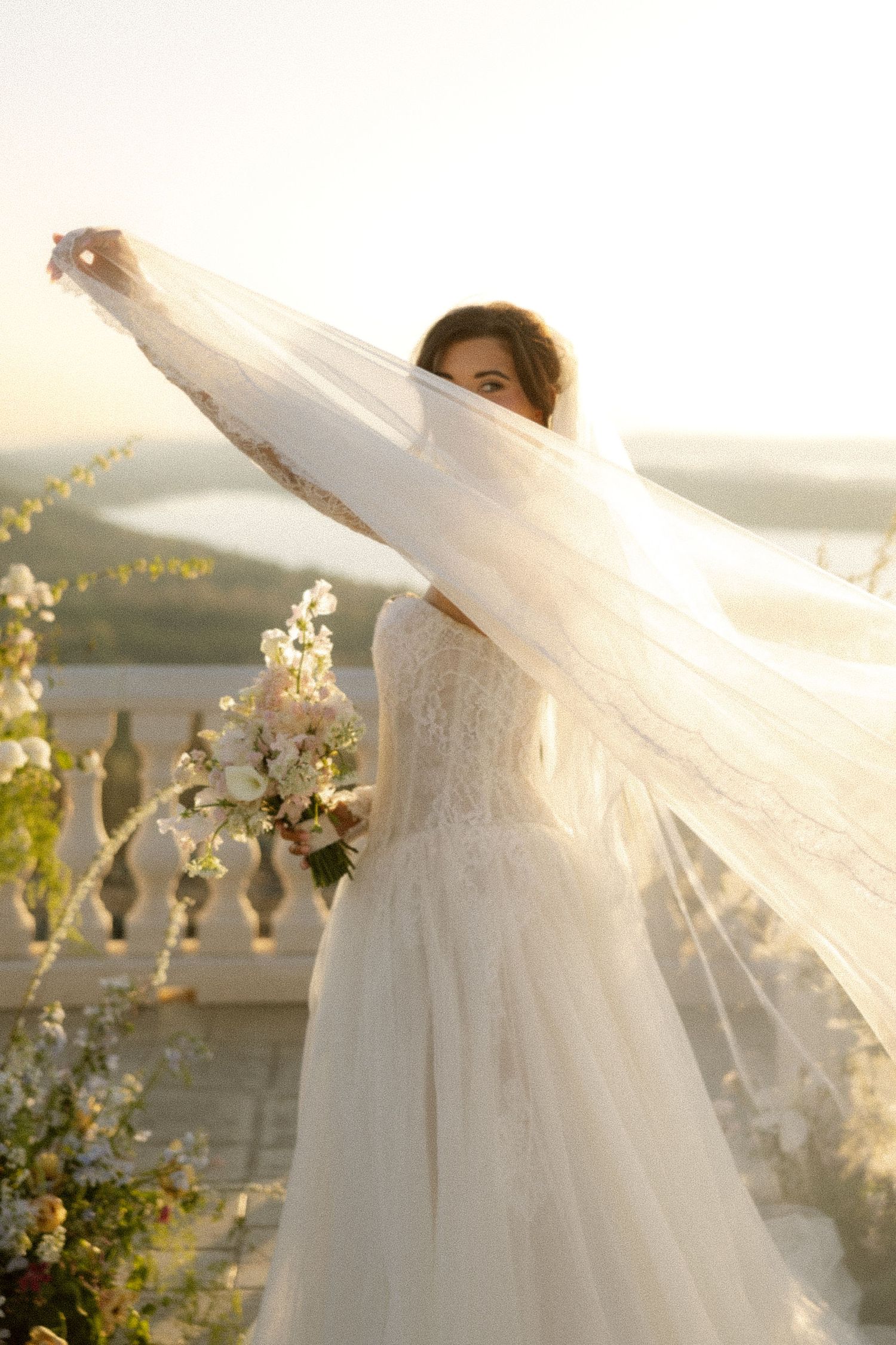 Bride in white lace gown holds flowing veil and bouquet on sunlit balcony with scenic landscape backdrop.